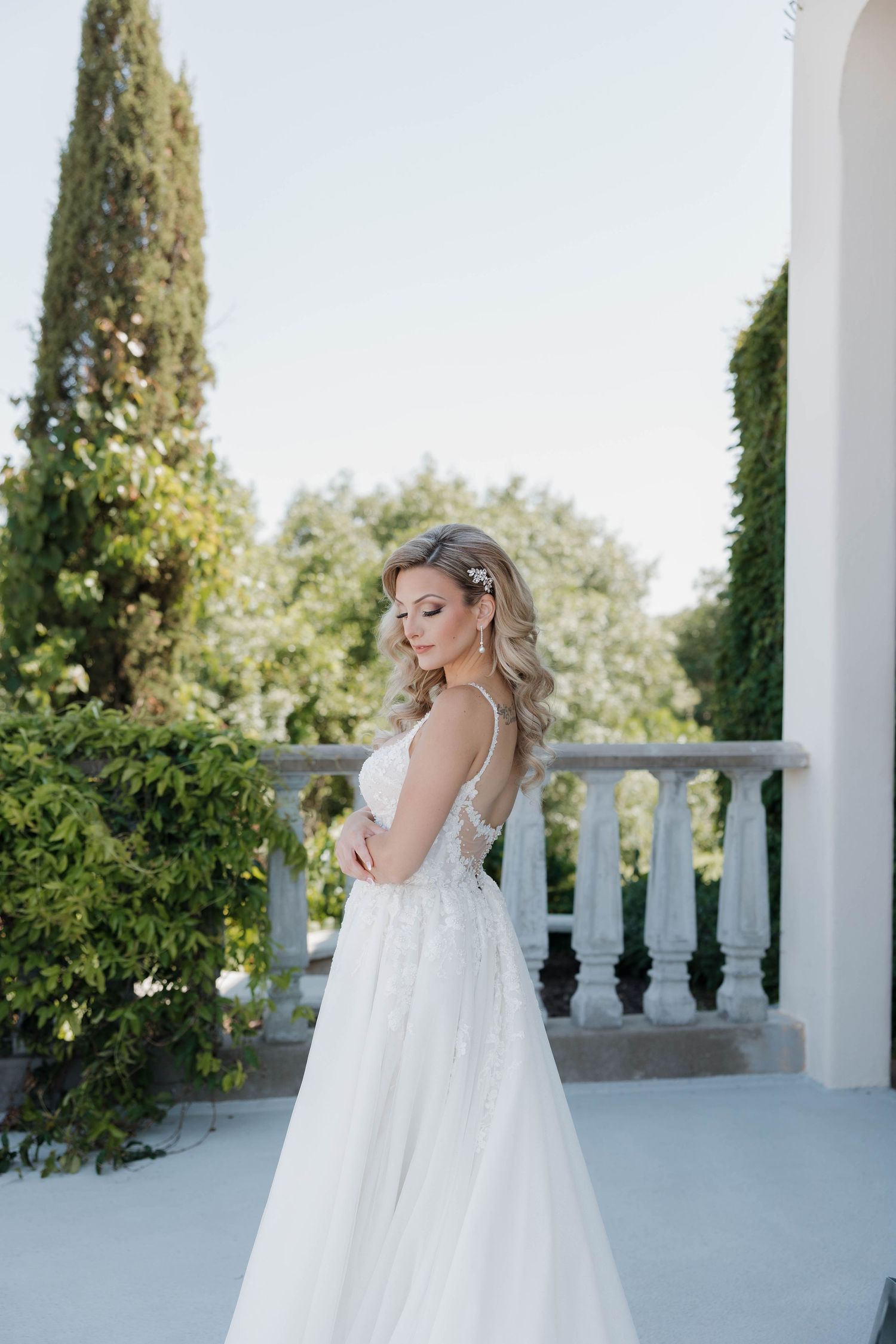Bride in white wedding dress poses elegantly on an outdoor terrace with white columns and green cypress trees.