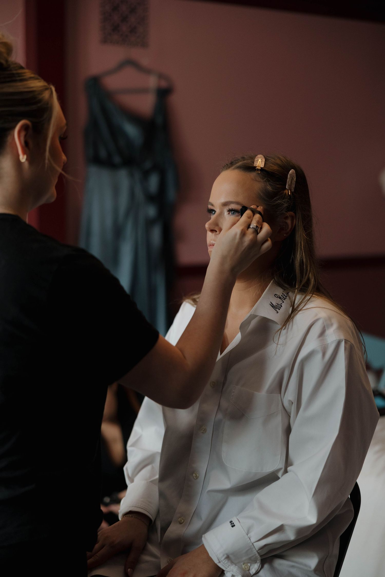 Makeup artist applies cosmetics to a client wearing a white robe in a dimly lit room during a beauty session.