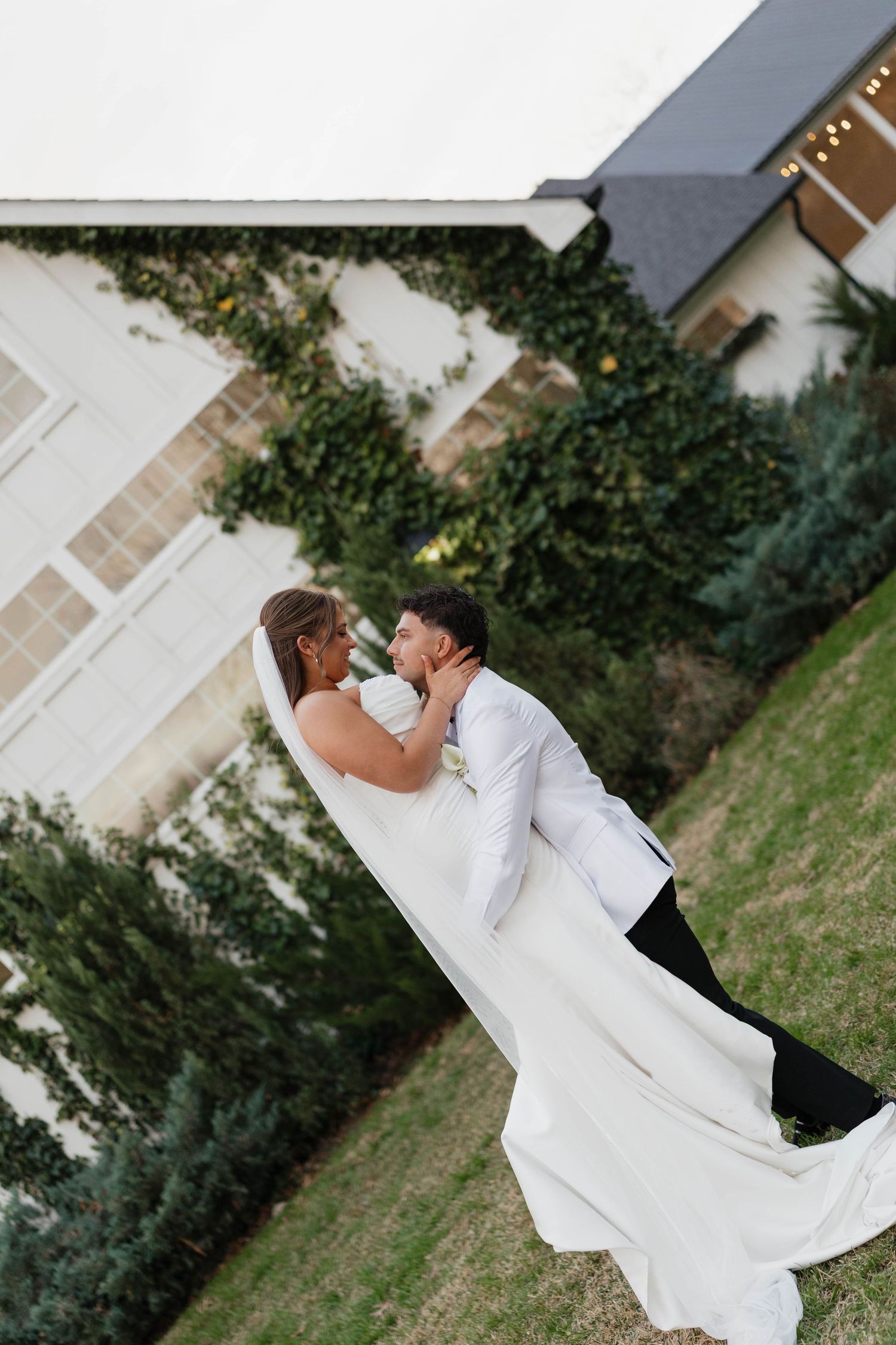 A couple shares a romantic moment outdoors, the bride in a flowing white gown and groom in a white suit near an ivy-covered building.