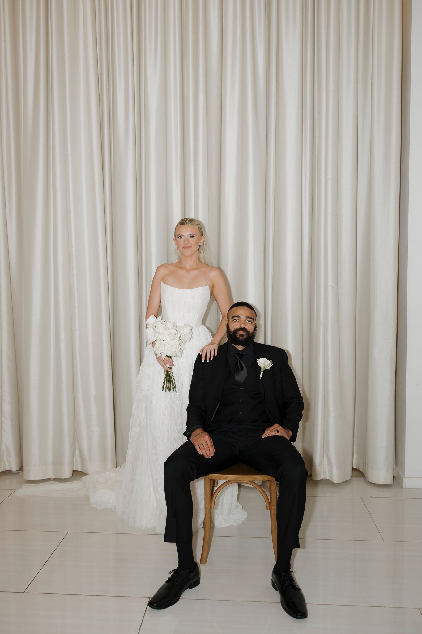 A bride in white dress sits next to a groom in black suit against a light curtain backdrop in three similar poses.