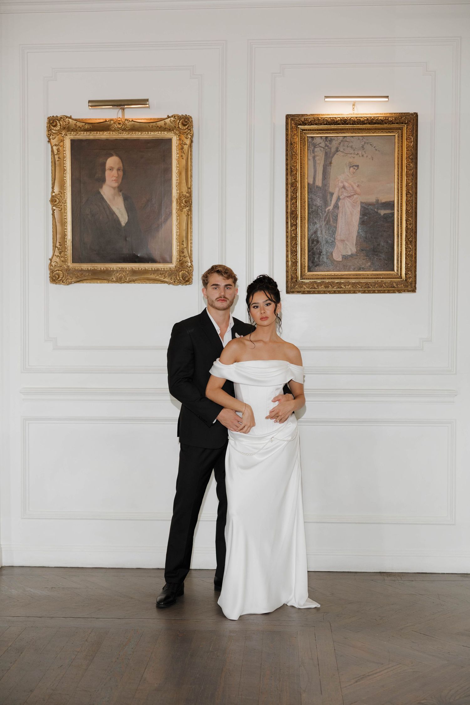 A romantic couple in wedding attire dance together in an elegant white room with ornate gold-framed paintings on the wall.