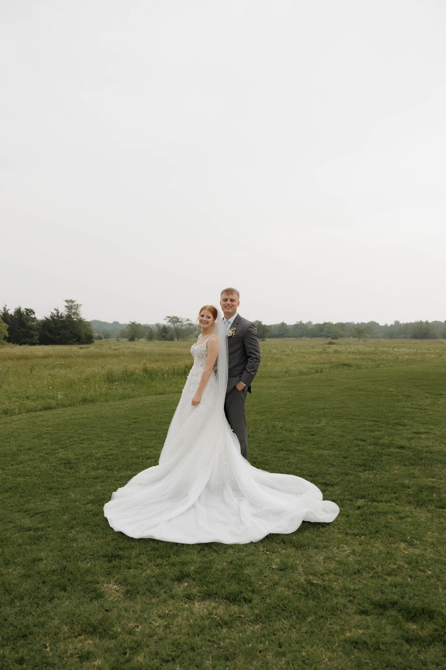 A couple stands together in formal wedding attire against a serene green meadow landscape at sunset.