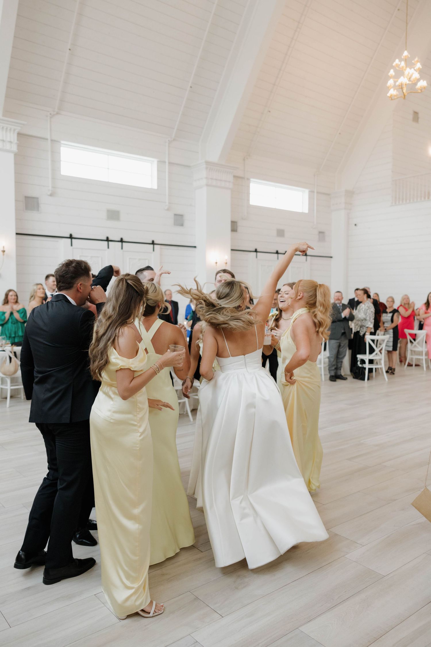 Wedding party celebrates together on a light wooden dance floor in a bright white venue.
