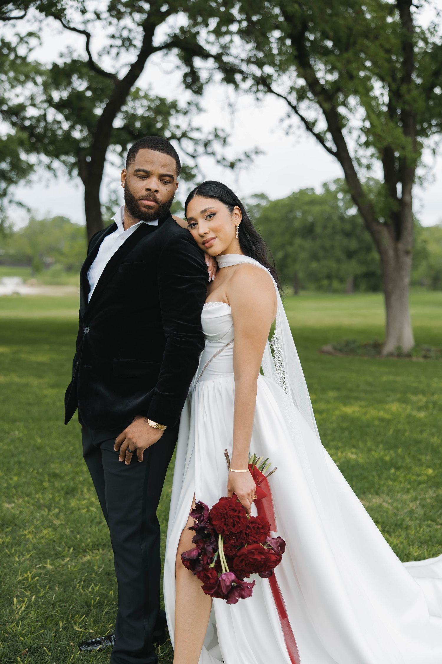 Elegant wedding couple in formal attire pose together on green lawn with bride holding burgundy bouquet.
