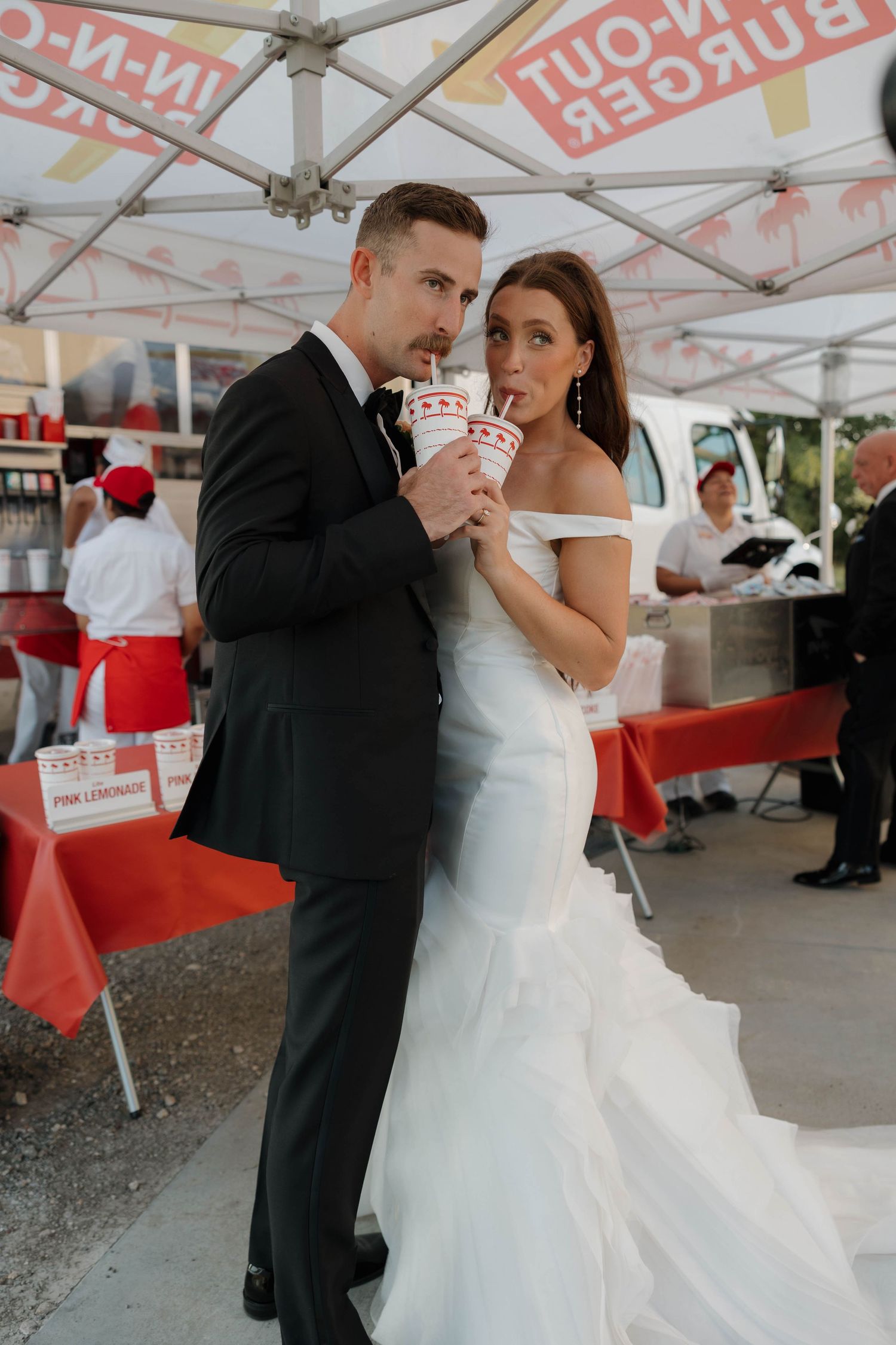 Couple enjoying In-N-Out Burger drinks under white tent at wedding reception.