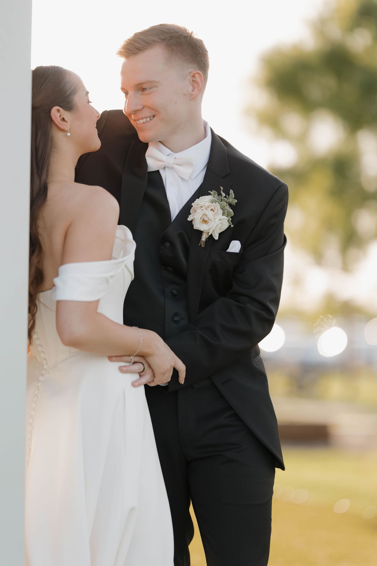 A bride in an off-shoulder white dress and groom in black tuxedo share an intimate moment outdoors at sunset.