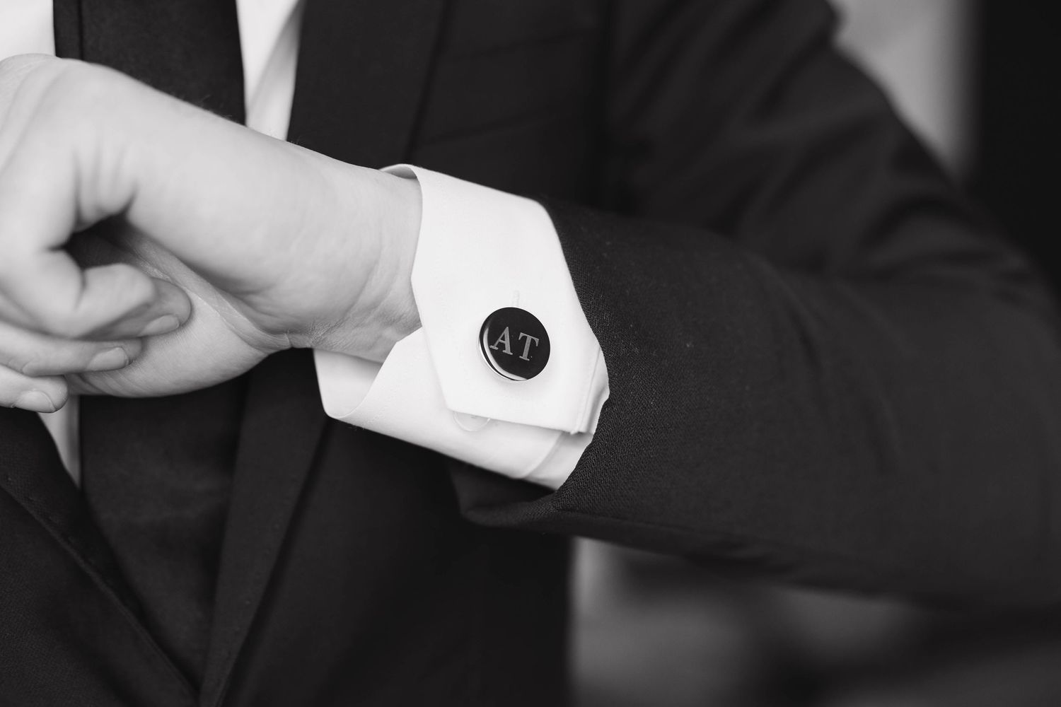 Close-up black and white photo of a person adjusting a formal black cufflink on a crisp white shirt cuff.