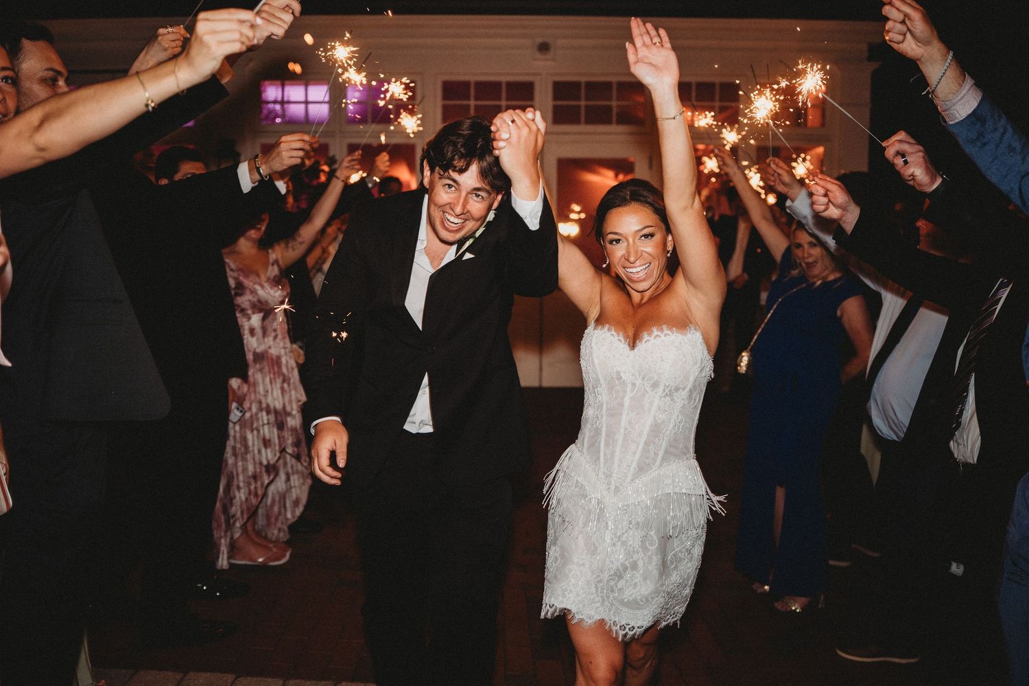 Bride and groom exit wedding reception through sparkler tunnel, guests cheering with raised arms in elegant venue.