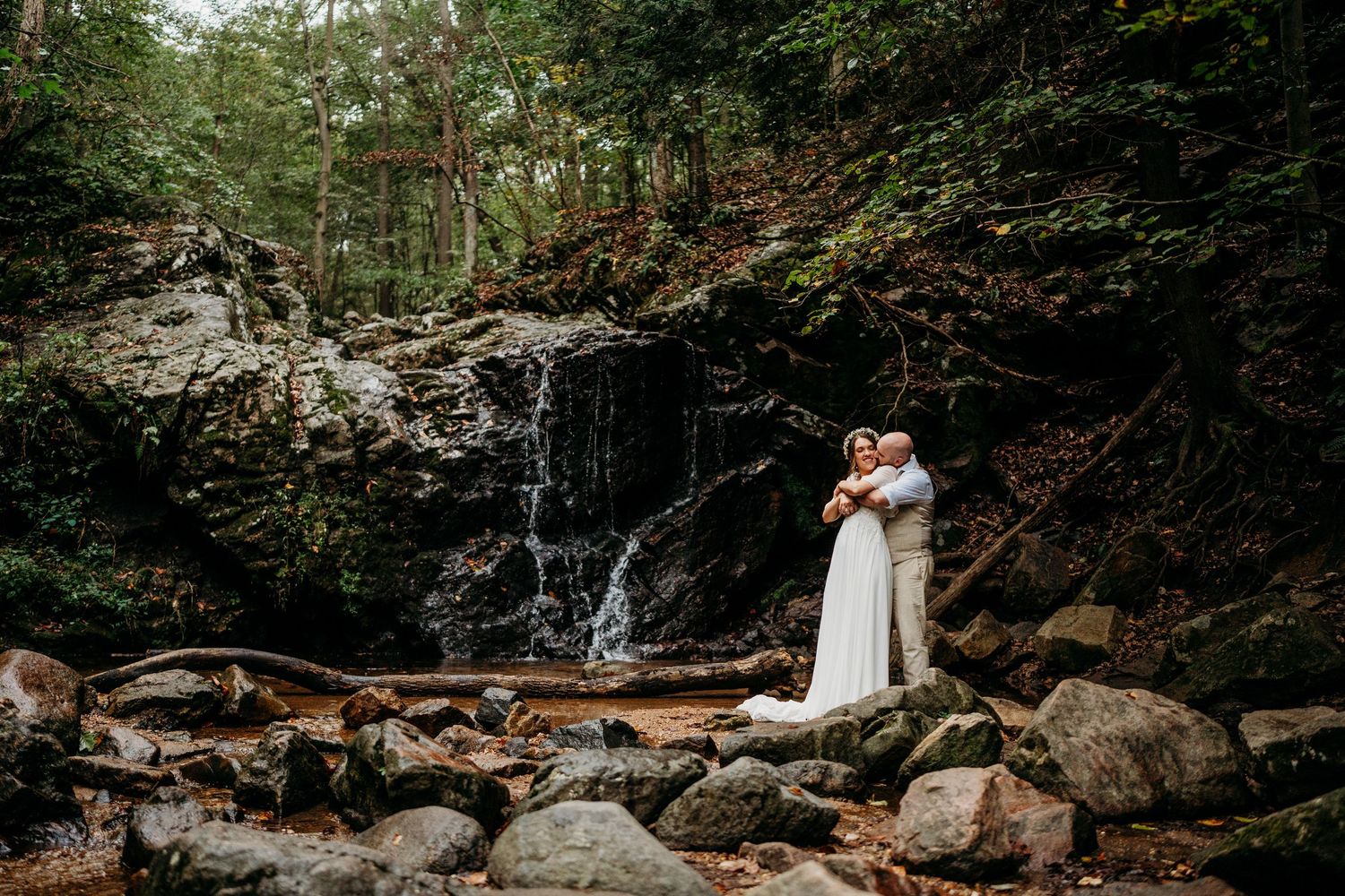 A couple shares a romantic moment by a cascading waterfall surrounded by lush forest and rocky terrain.