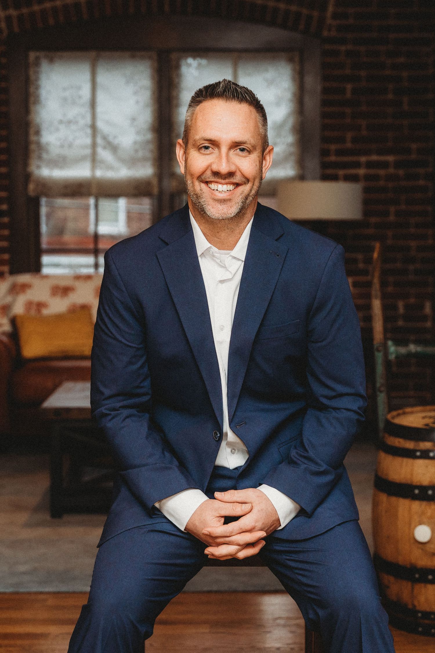 A professional headshot of a person in a navy suit seated in an industrial-style room.