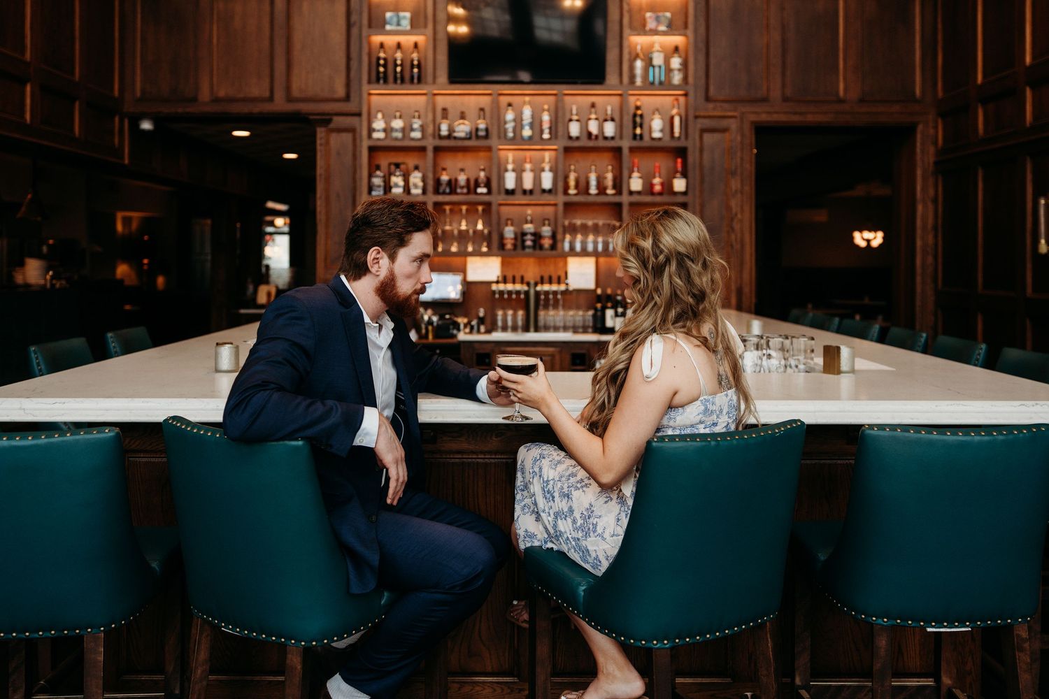 People sitting at a dimly lit bar counter with bottles and glasses illuminated by warm ambient lighting.