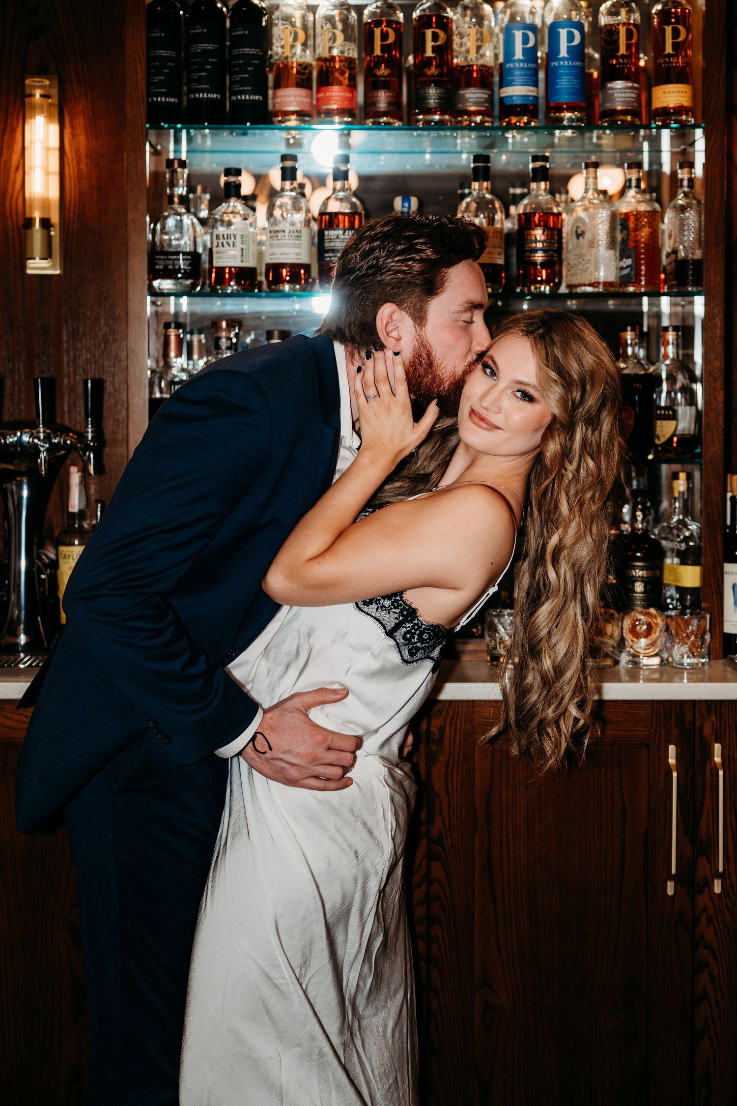 A romantic couple shares a passionate kiss at a dimly lit bar with shelves of bottles in the background.