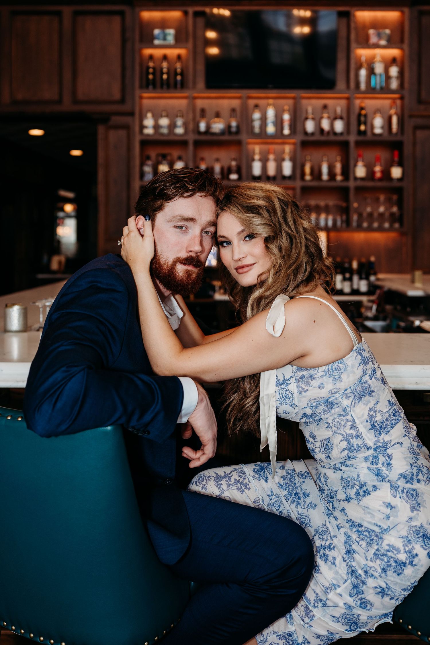 Romantic couple in blue dress and suit share intimate moments at an elegant bar with bottles displayed in the background.