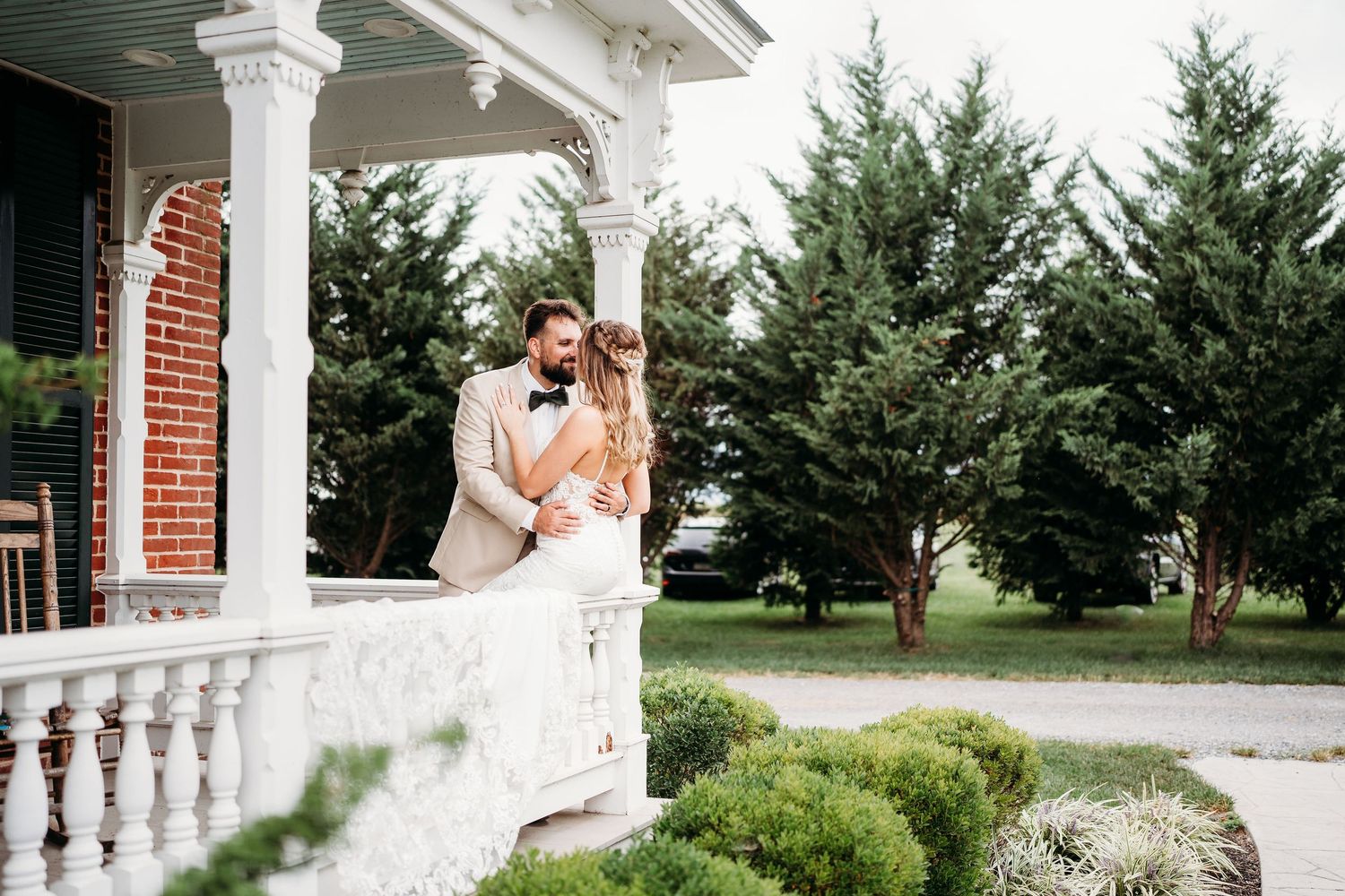 An elegant couple shares a romantic moment on a classic white columned porch with evergreen trees in the background.