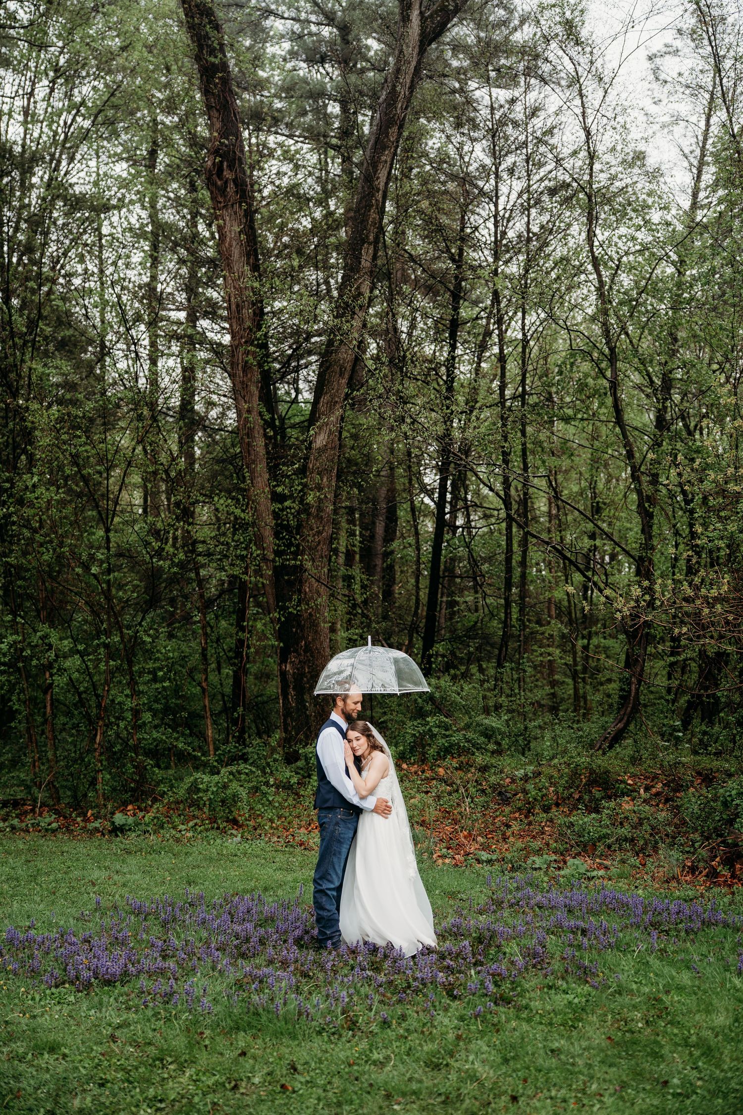 Bride and groom embrace under a clear umbrella in a lush forest clearing with purple wildflowers on a rainy wedding day.