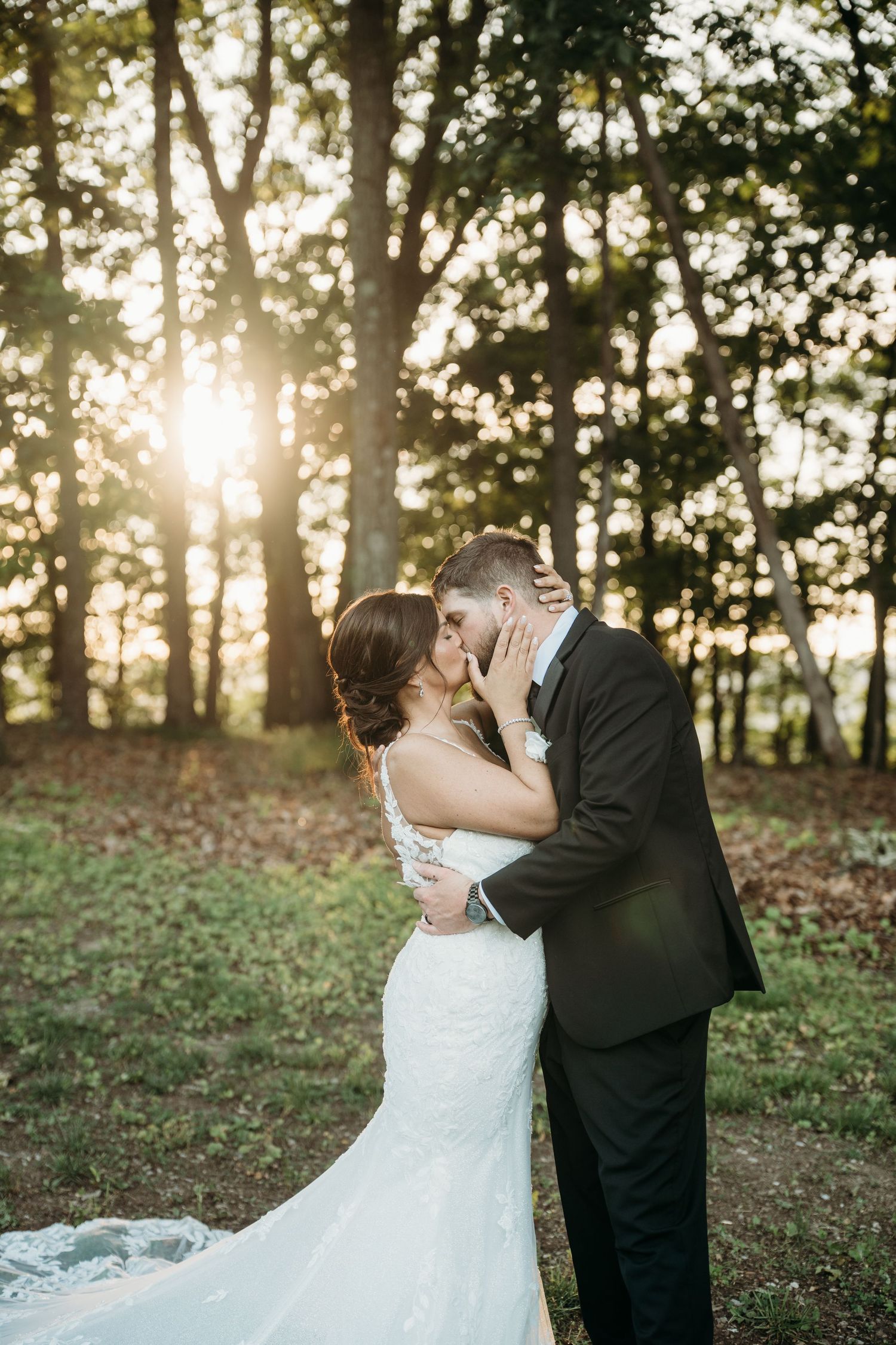 Bride and groom share a romantic kiss in a sunlit forest, bride wearing a lace mermaid gown with a flowing train.
