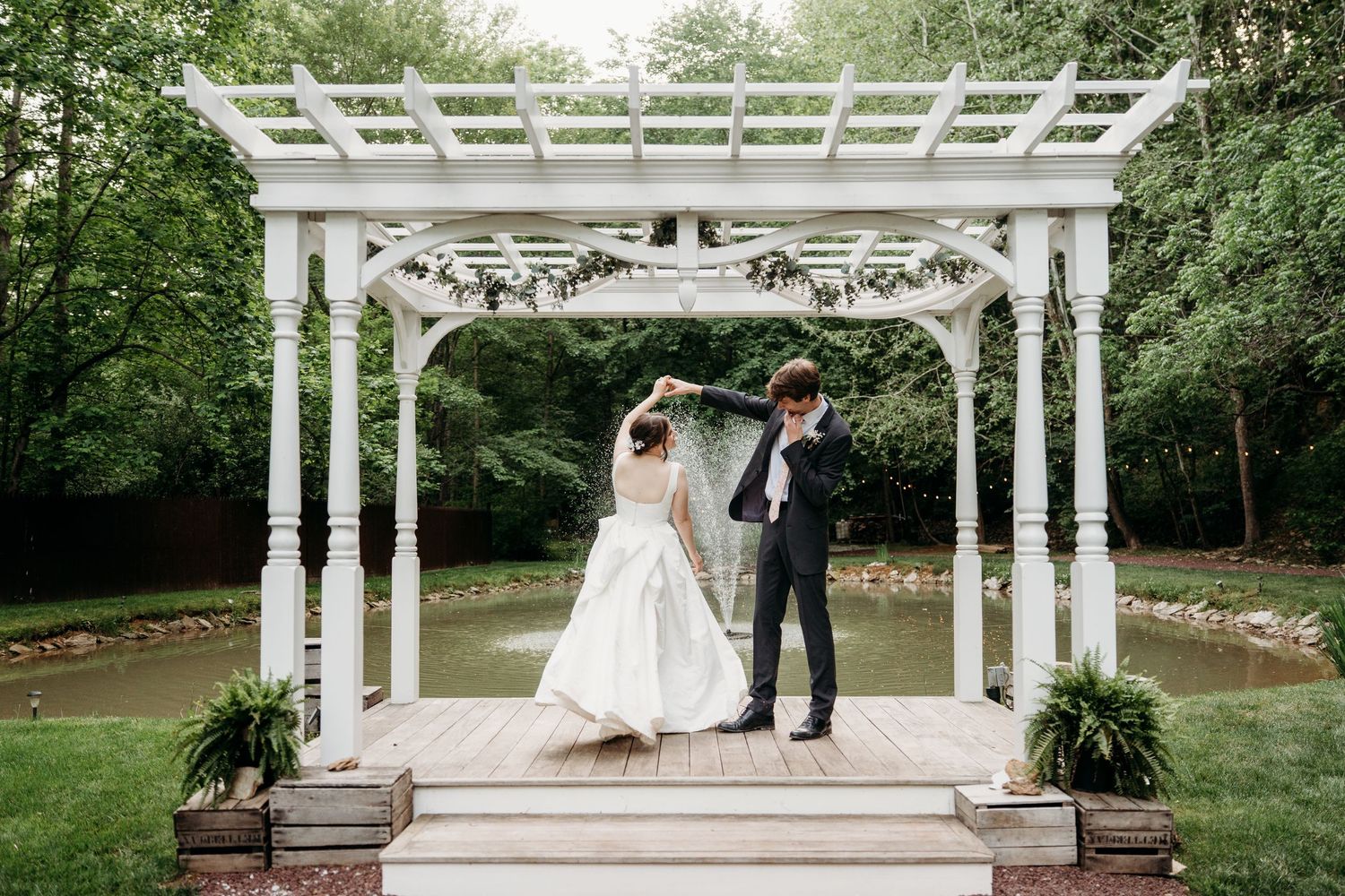 Bride and groom stand under white pergola on wooden deck surrounded by lush greenery near a pond.