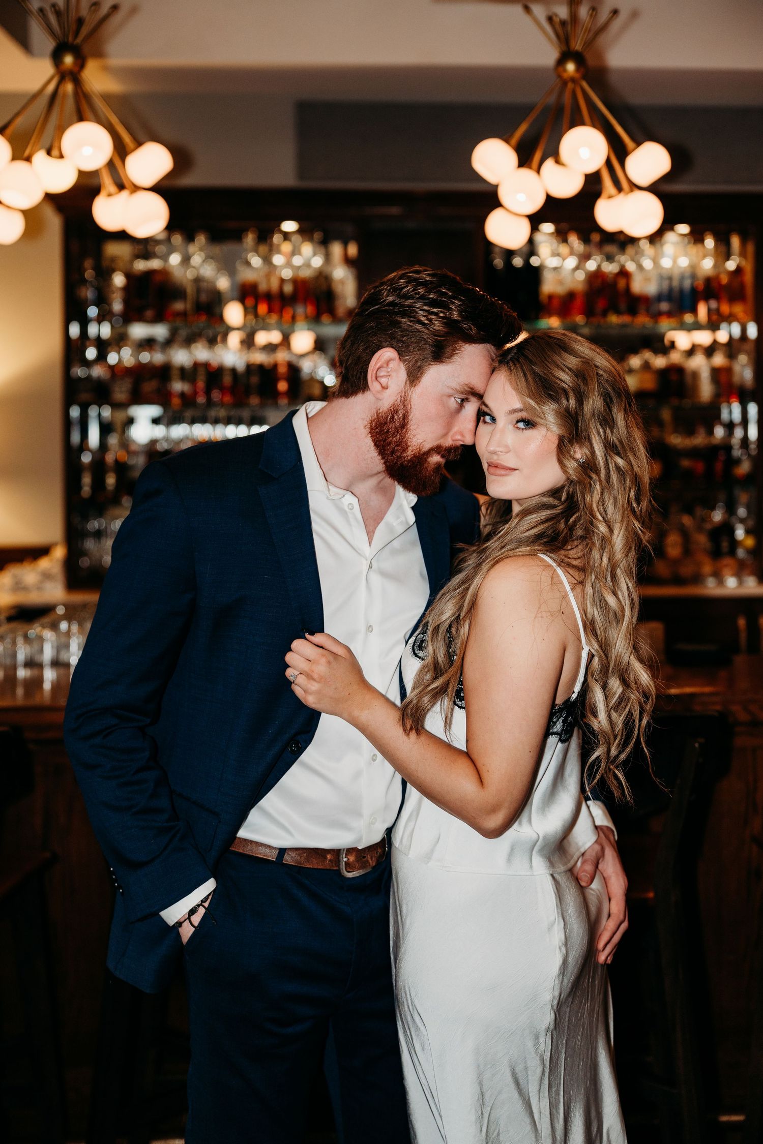 Romantic couple embraces in elegant attire at dimly lit bar with glowing pendant lights overhead.