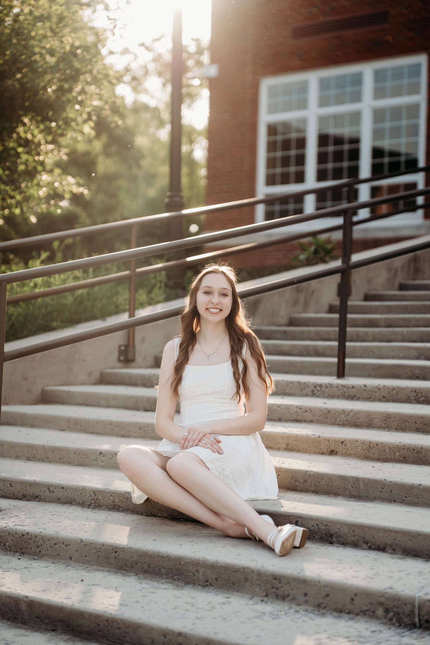 Young woman in white dress sitting on outdoor steps, smiling in golden hour sunlight near a brick building.