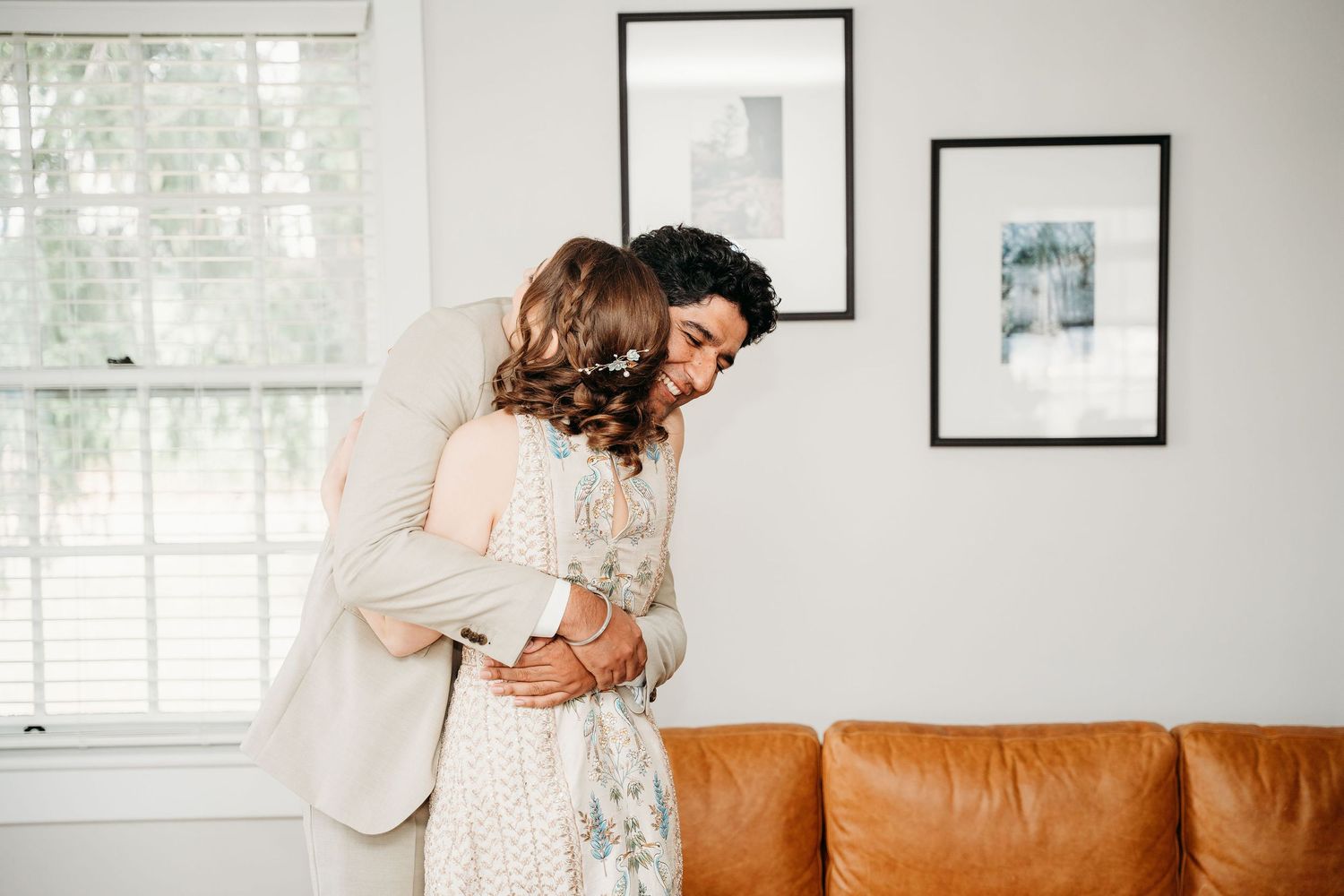A tender embrace between two people in front of a tan leather couch and framed artwork on a white wall.