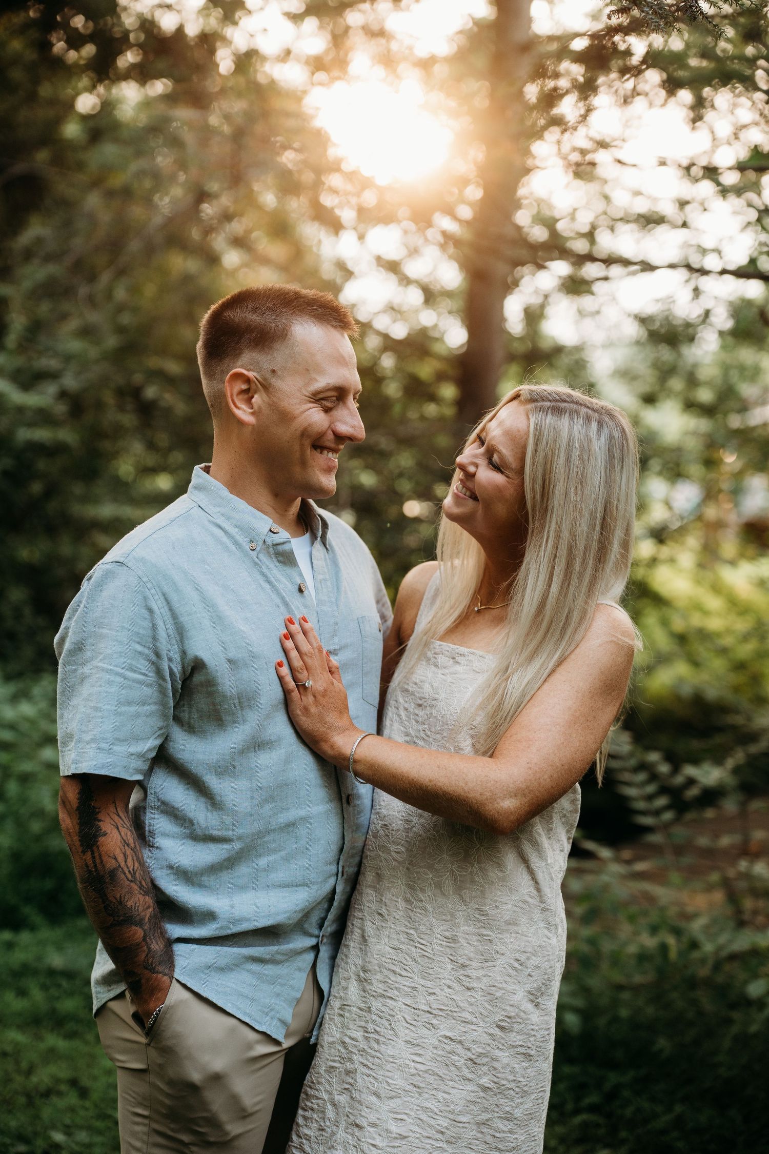 Couple smiling at each other in a sunlit forest, man in blue shirt with tattoos, woman in white dress.