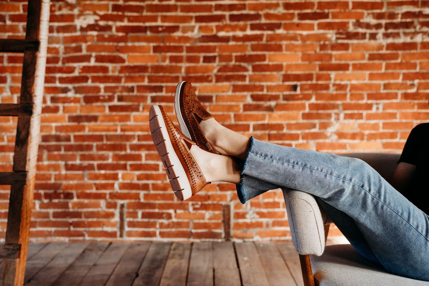Two people sit and chat on a wooden bench against a rustic red brick wall in a casual indoor setting.