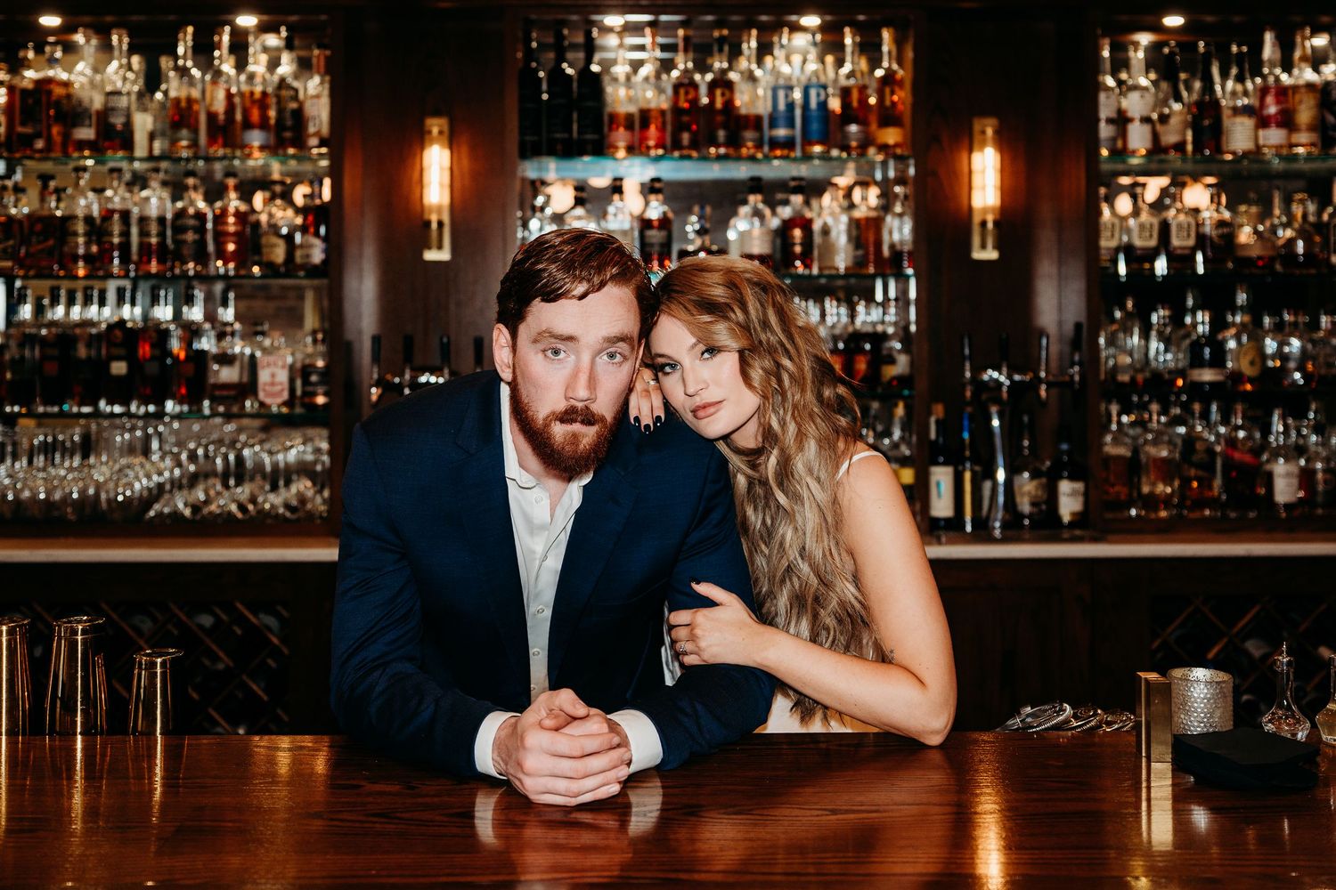 A couple embraces intimately at a dimly lit bar with bottles and glassware displayed on shelves in the background.