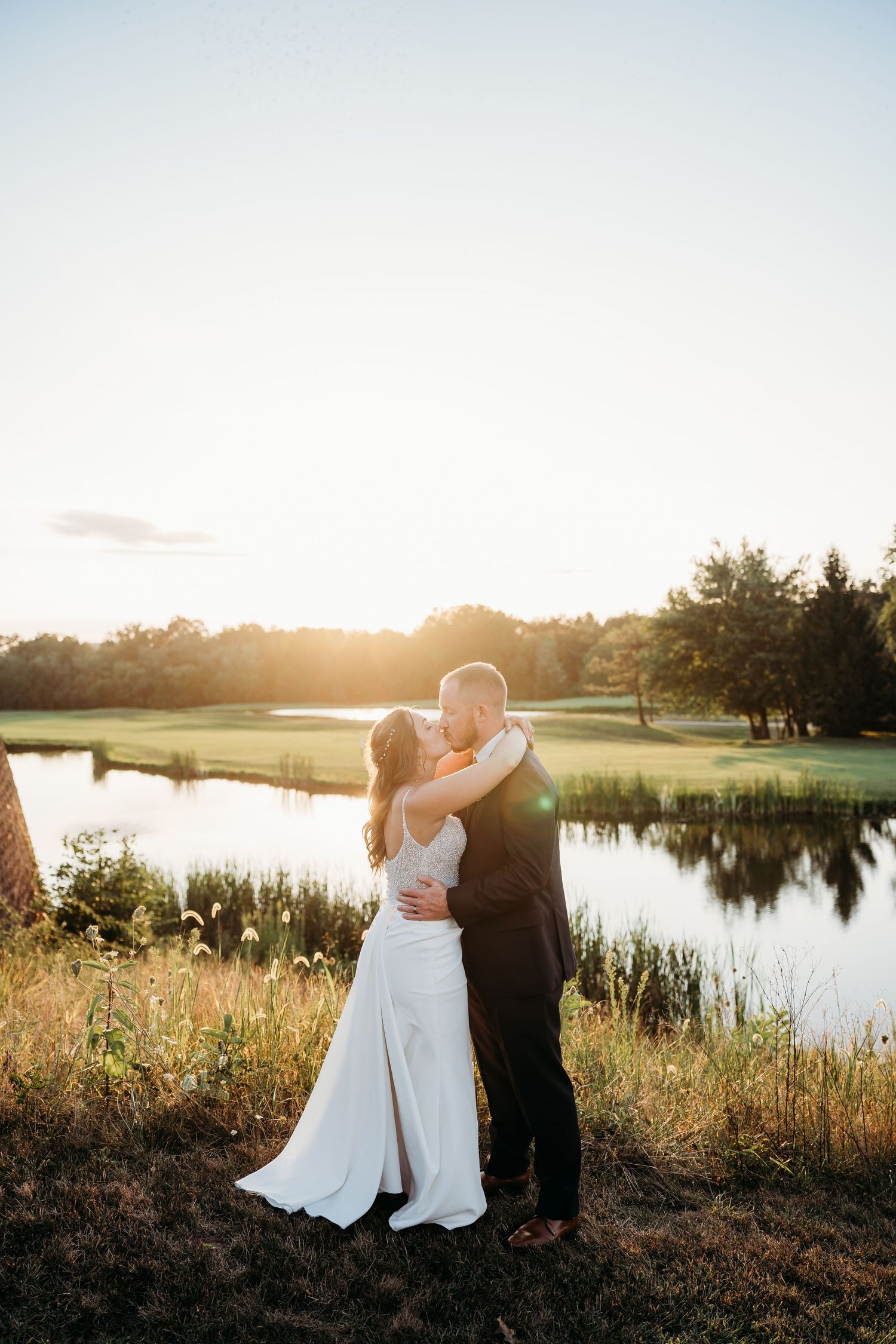 A couple embraces during golden hour sunset by a serene pond on a golf course with tall grass in the foreground.
