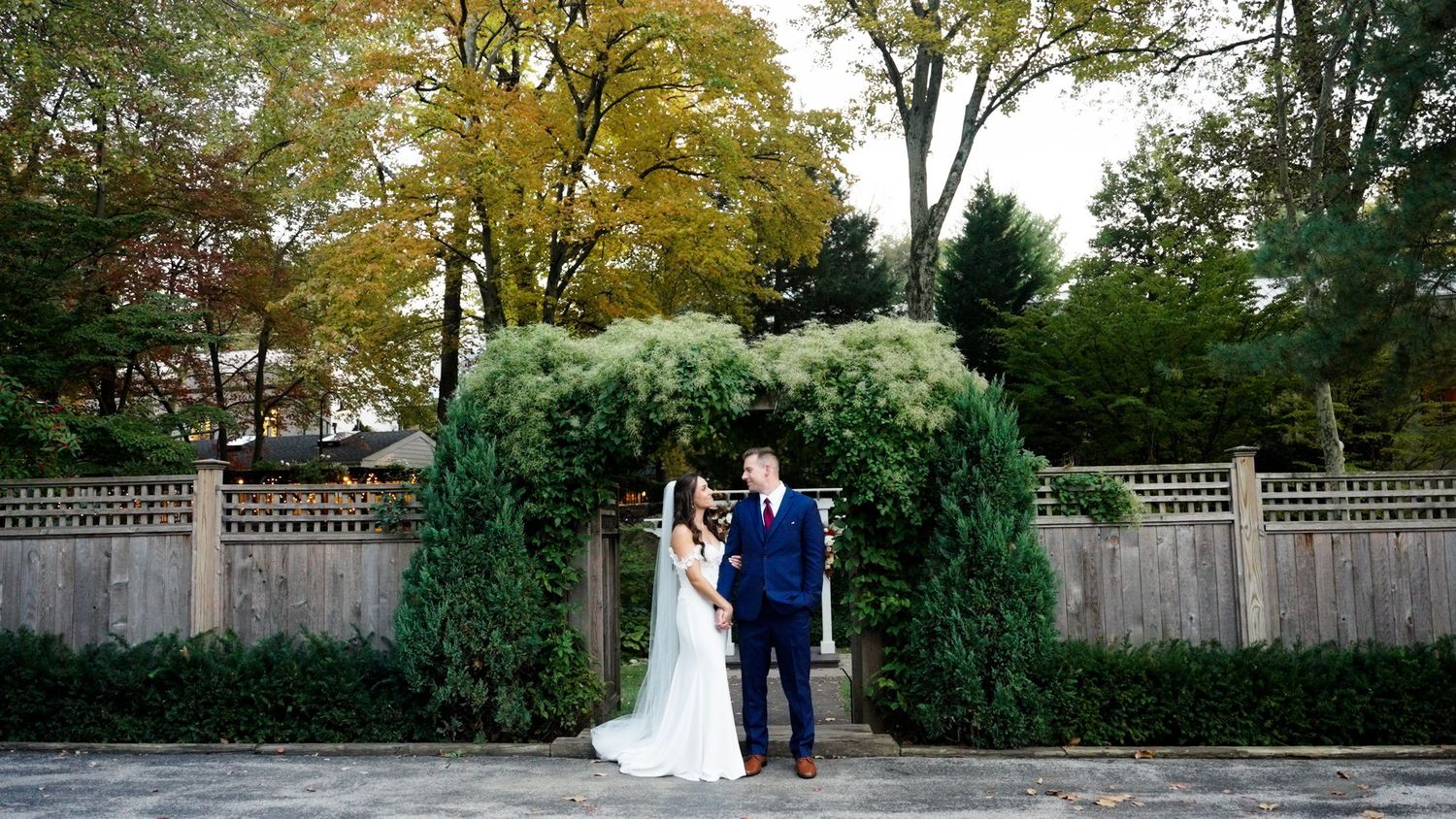 Wedding couple poses under green garden arch with fall foliage in background.