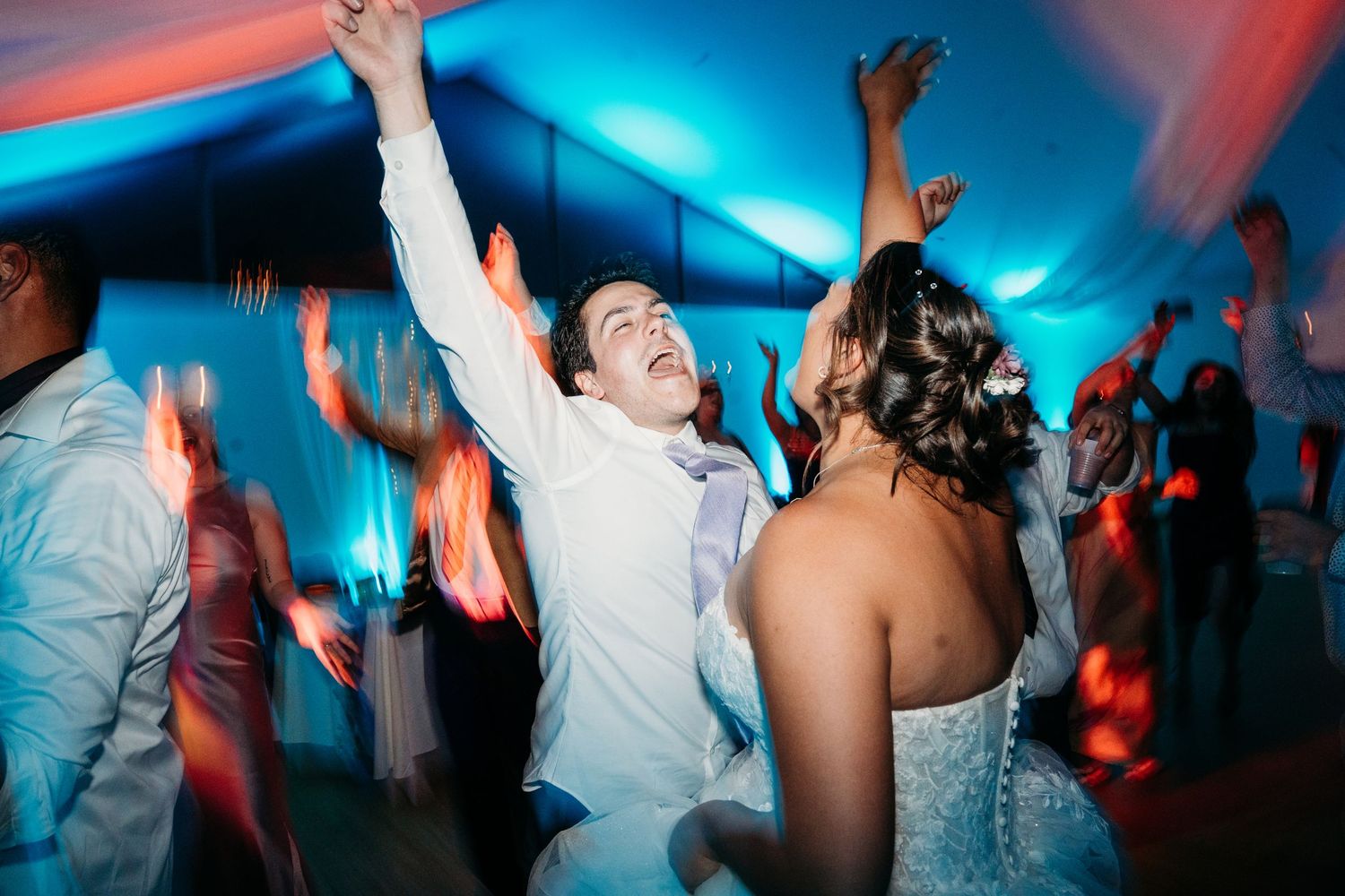 Wedding guests dance with raised arms under vibrant red and blue lighting on a lively dance floor.