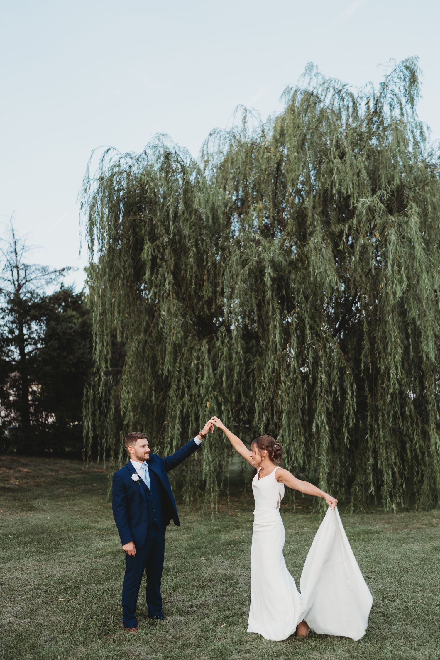 Bride and groom dancing outdoors under a weeping willow tree, groom in navy suit twirling bride in white gown.