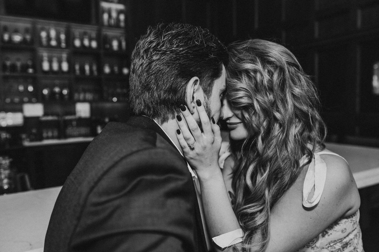 A black and white photo of a couple kissing while sitting on a bench in an urban setting.
