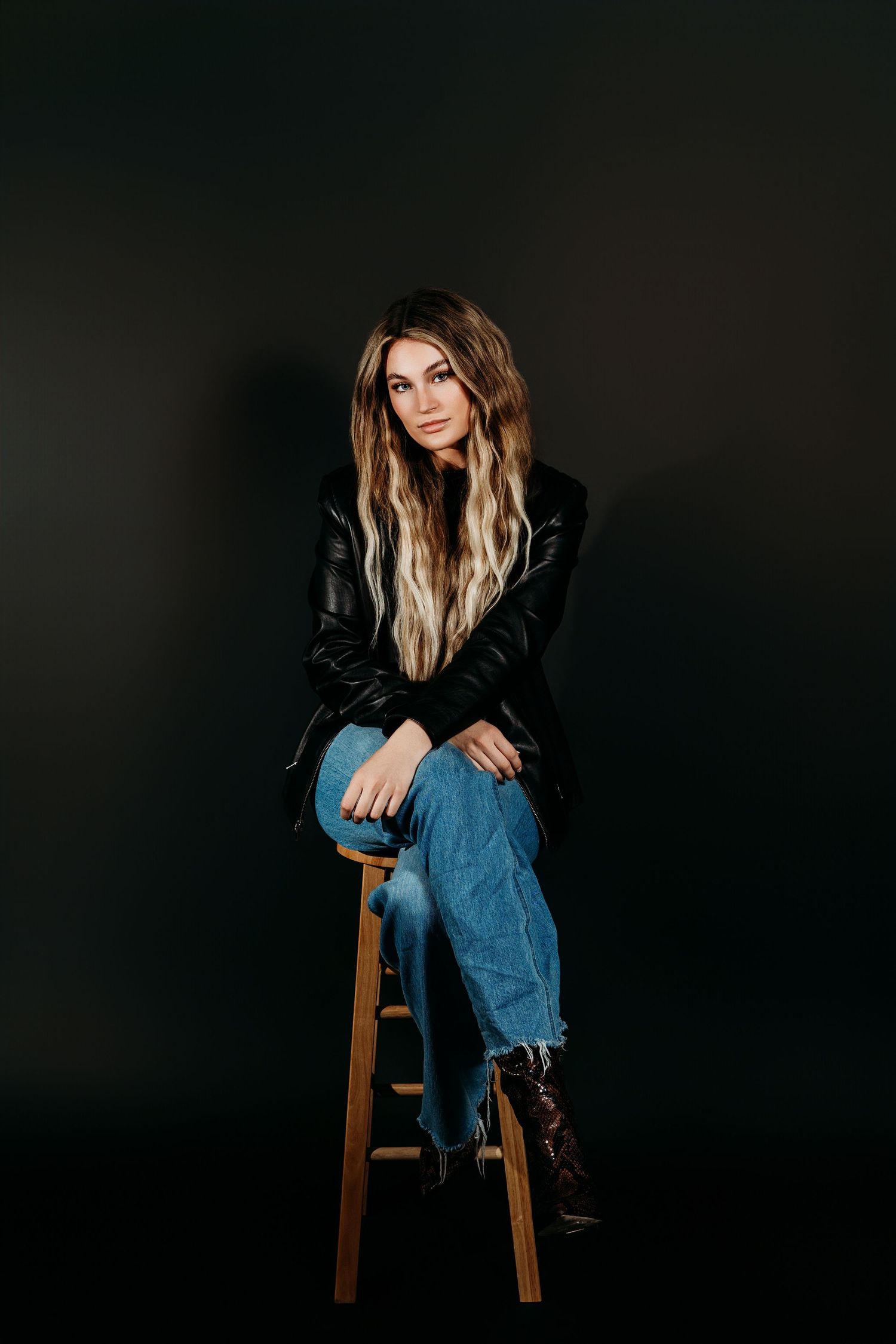 A model in black leather jacket and blue jeans poses on wooden stool against dark background in studio setting.