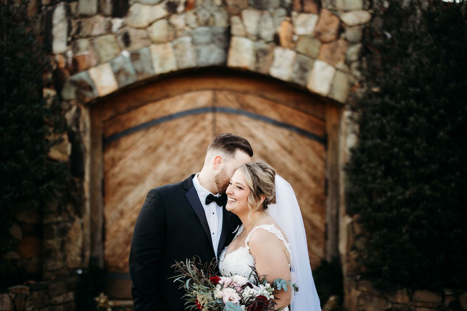 A romantic wedding couple embraces in front of a rustic stone archway with elegant floral bouquet.