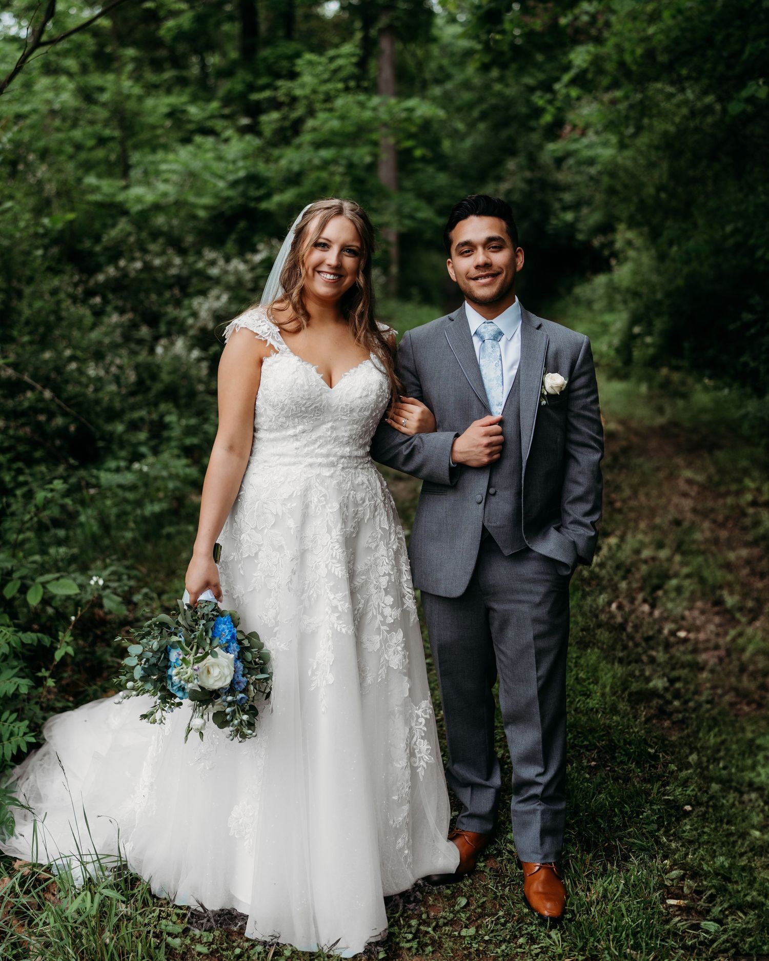 A couple stands together outdoors in wedding attire with lush greenery behind them and a blue and white bouquet.