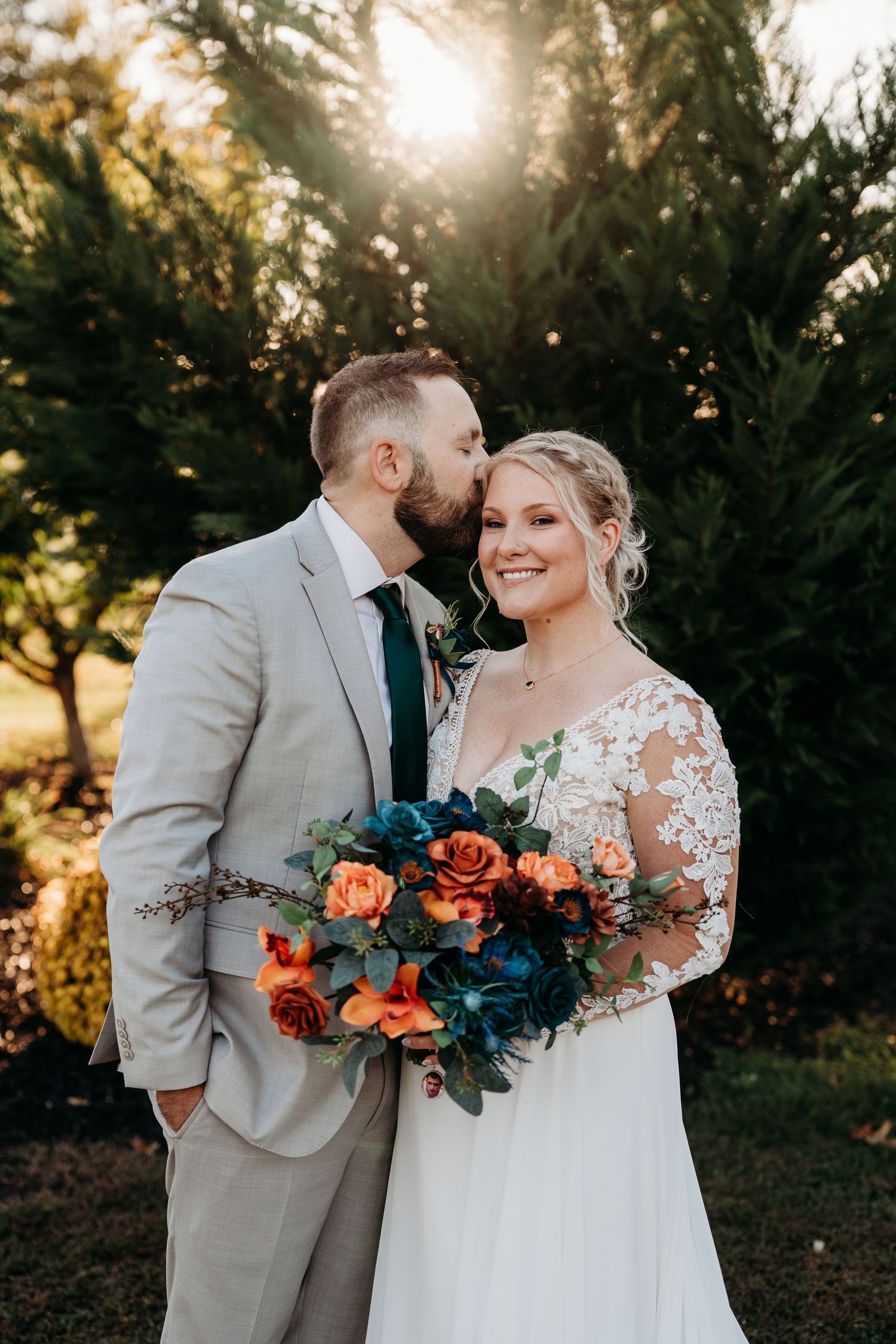 A joyful wedding couple poses outdoors with a vibrant orange and blue bouquet during golden hour.