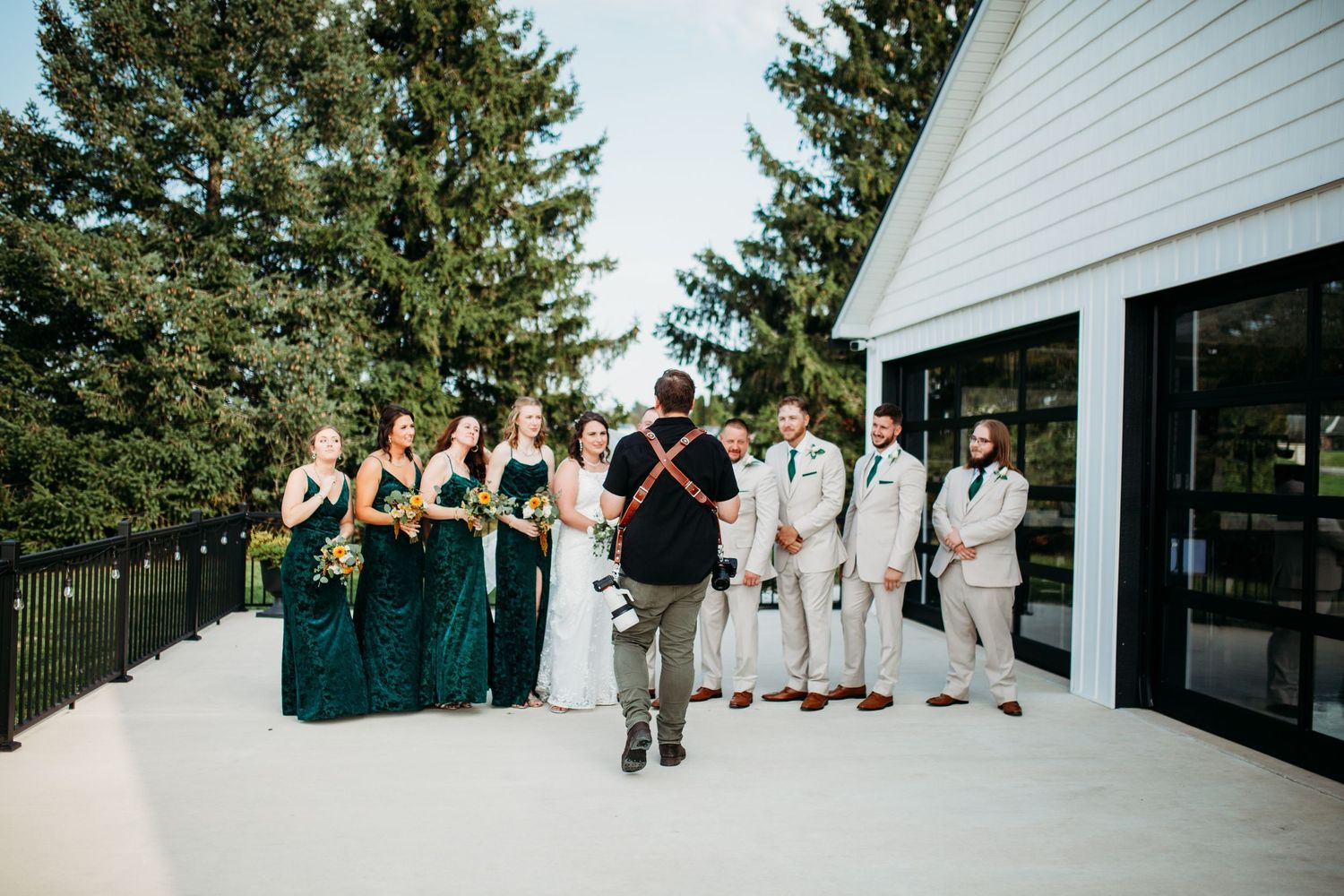 Wedding party poses together on a white patio deck with bridesmaids in emerald dresses and groomsmen in light suits.