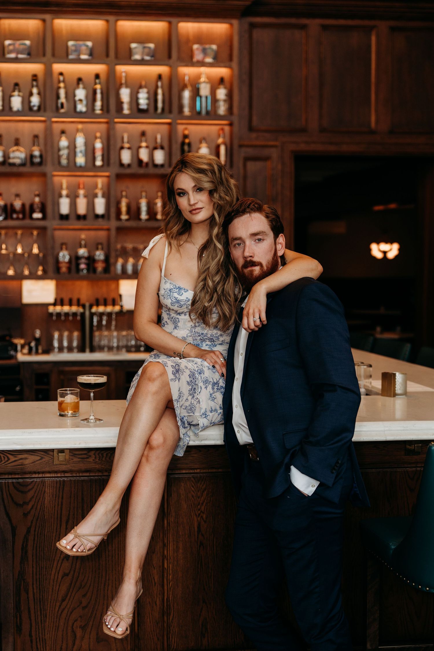 A couple poses intimately at an upscale bar with wooden decor and illuminated bottle shelves in the background.