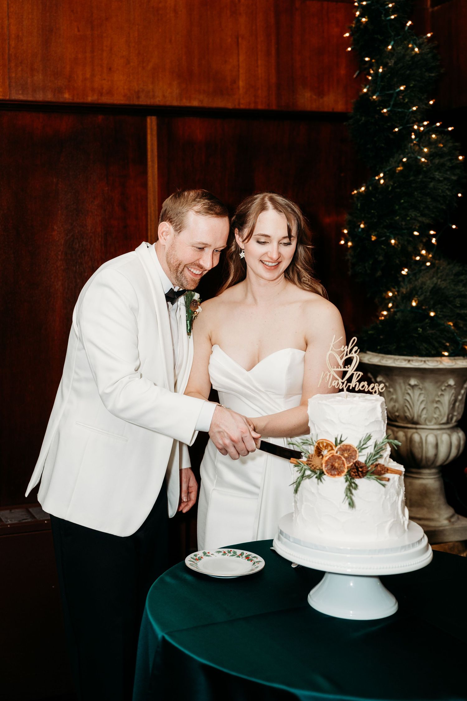 A couple cuts into their elegant white wedding cake together during their winter reception with twinkle lights in background.