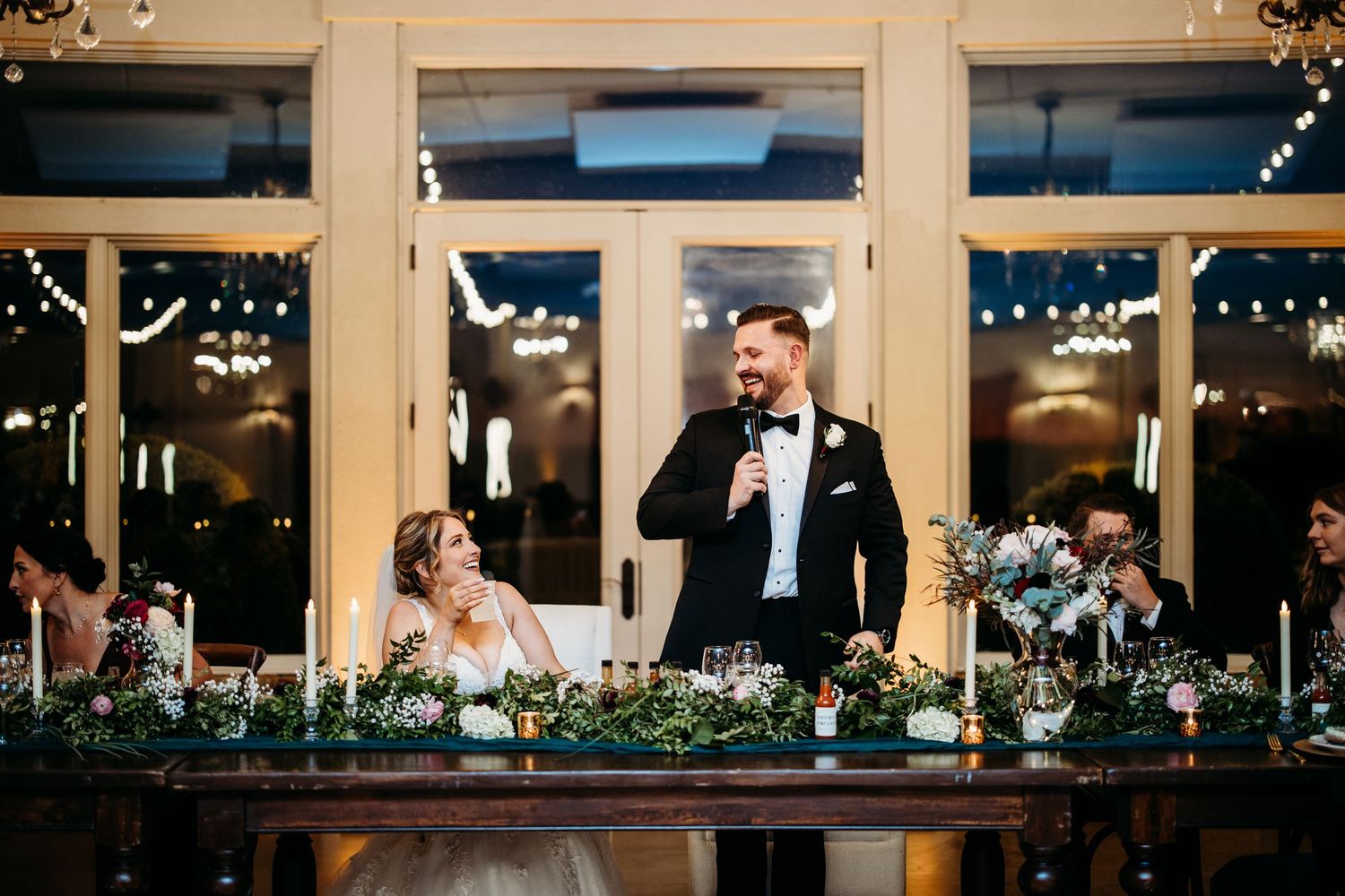 Groom speaks into microphone at wedding reception head table while bride smiles beside floral greenery centerpieces.
