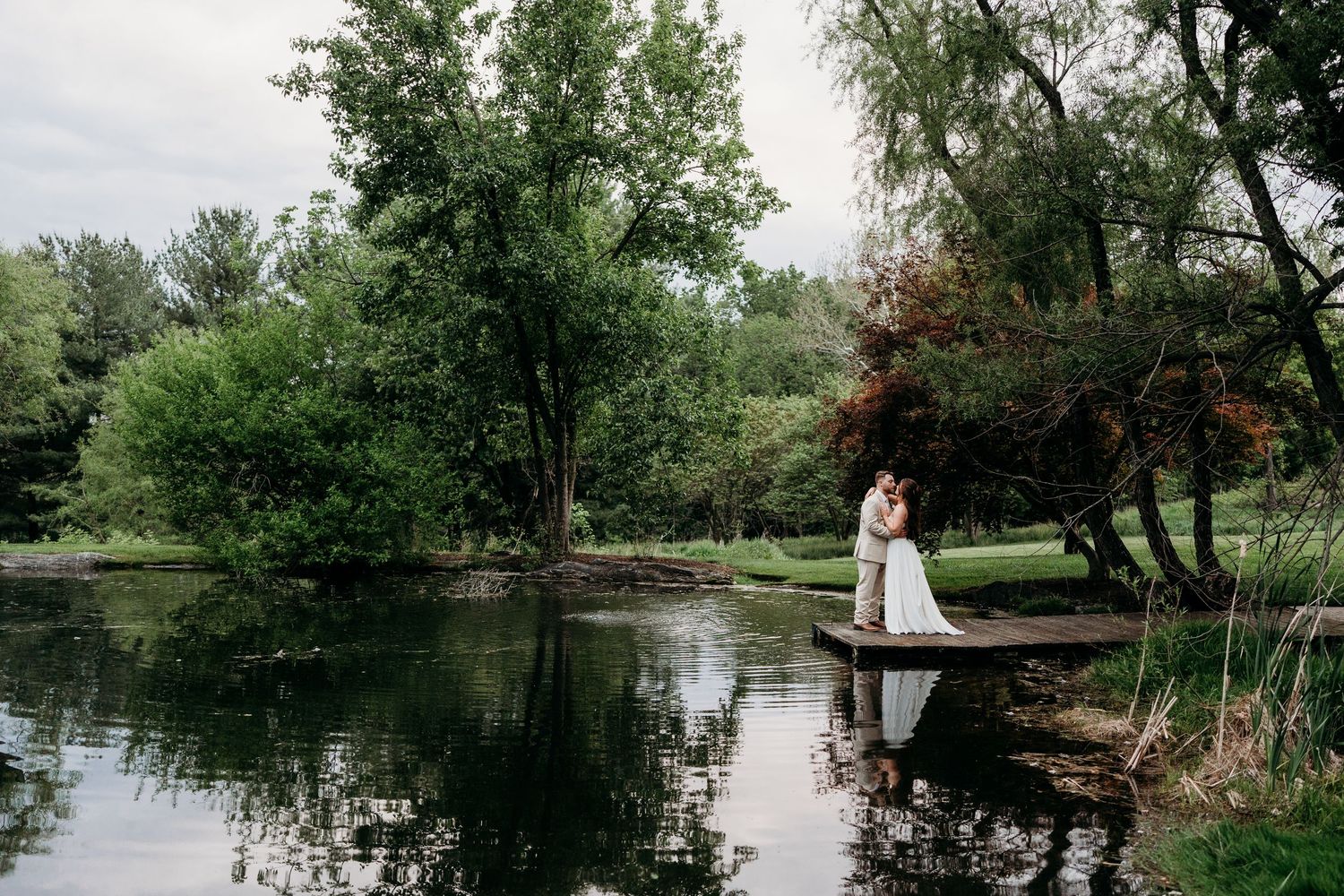 A wedding couple embraces on a wooden dock surrounded by a serene pond and lush green trees.