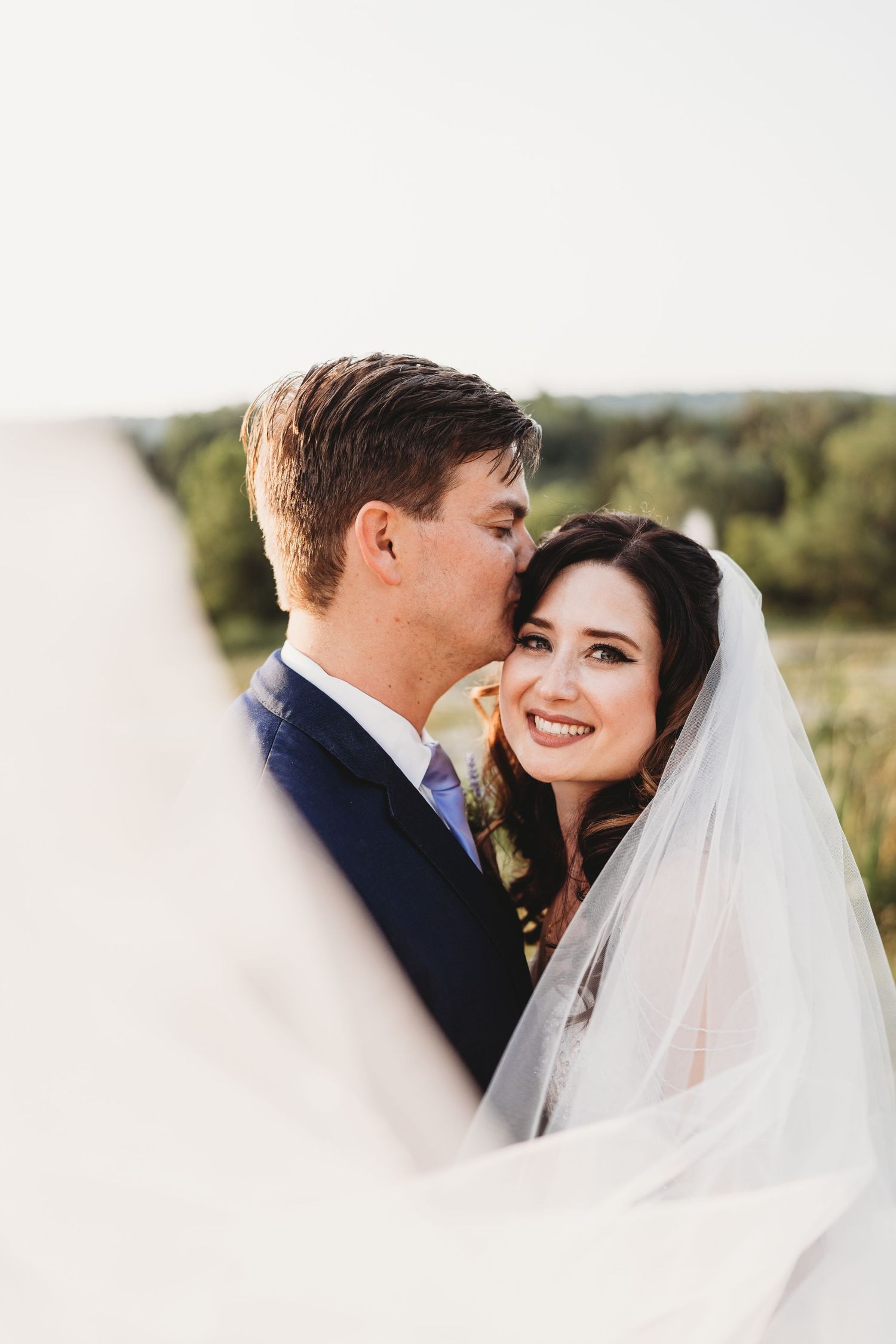 A romantic wedding portrait captured outdoors with a flowing white veil and natural landscape in the background.