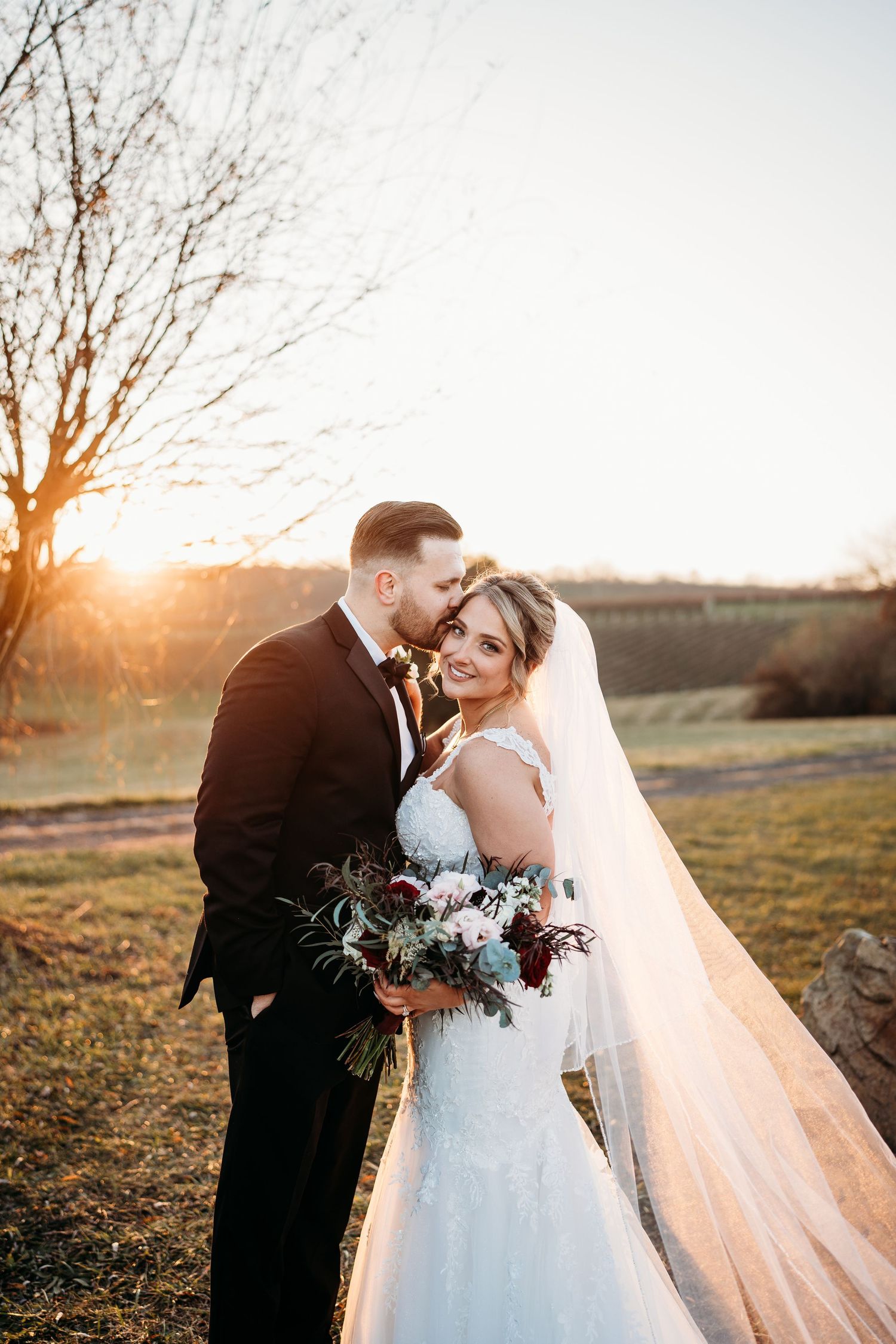 Newlyweds share intimate moment at sunset in outdoor wedding venue with long flowing veil and floral bouquet.