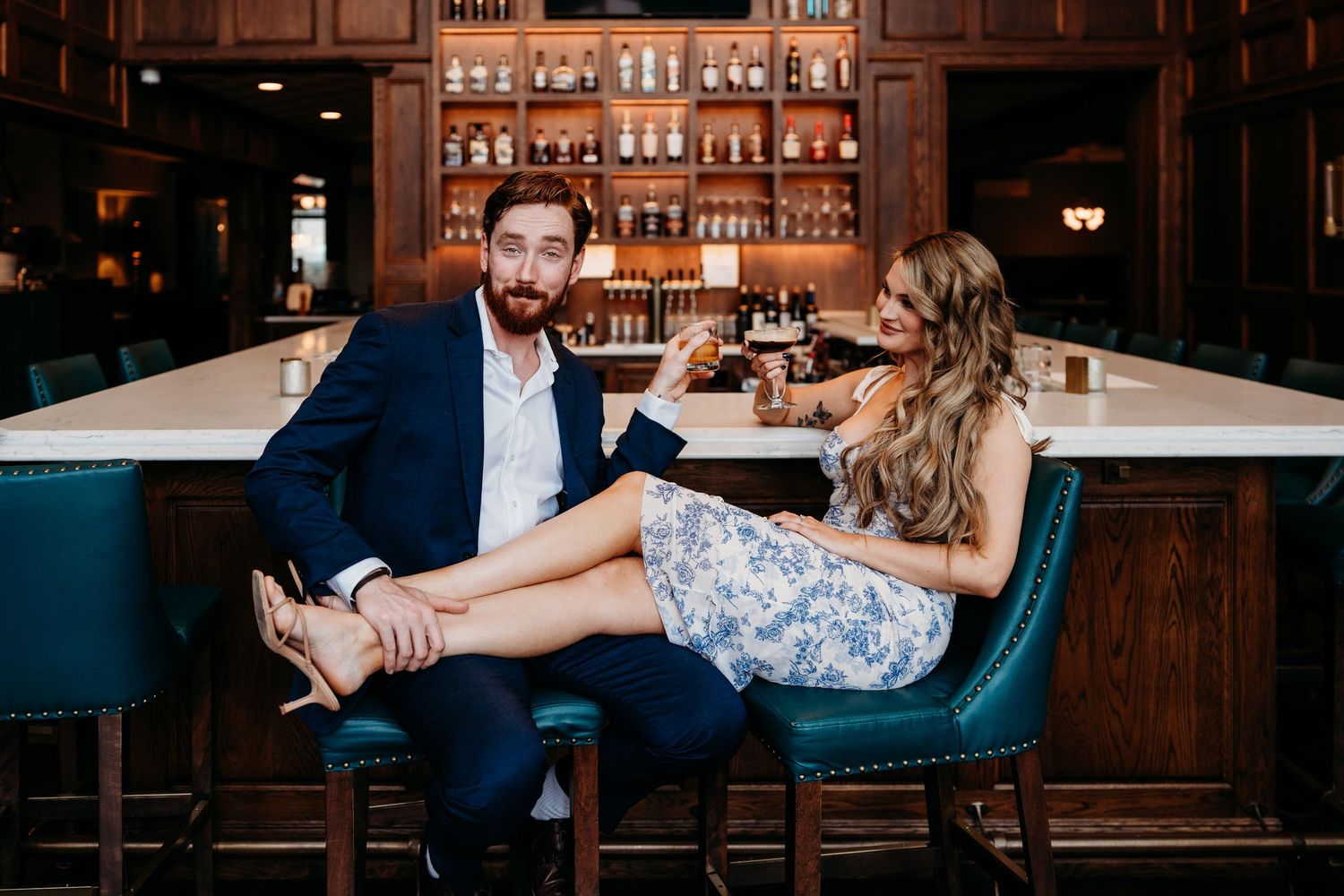 A romantic couple shares an intimate moment at an elegant bar with wooden shelving and warm lighting in the background.