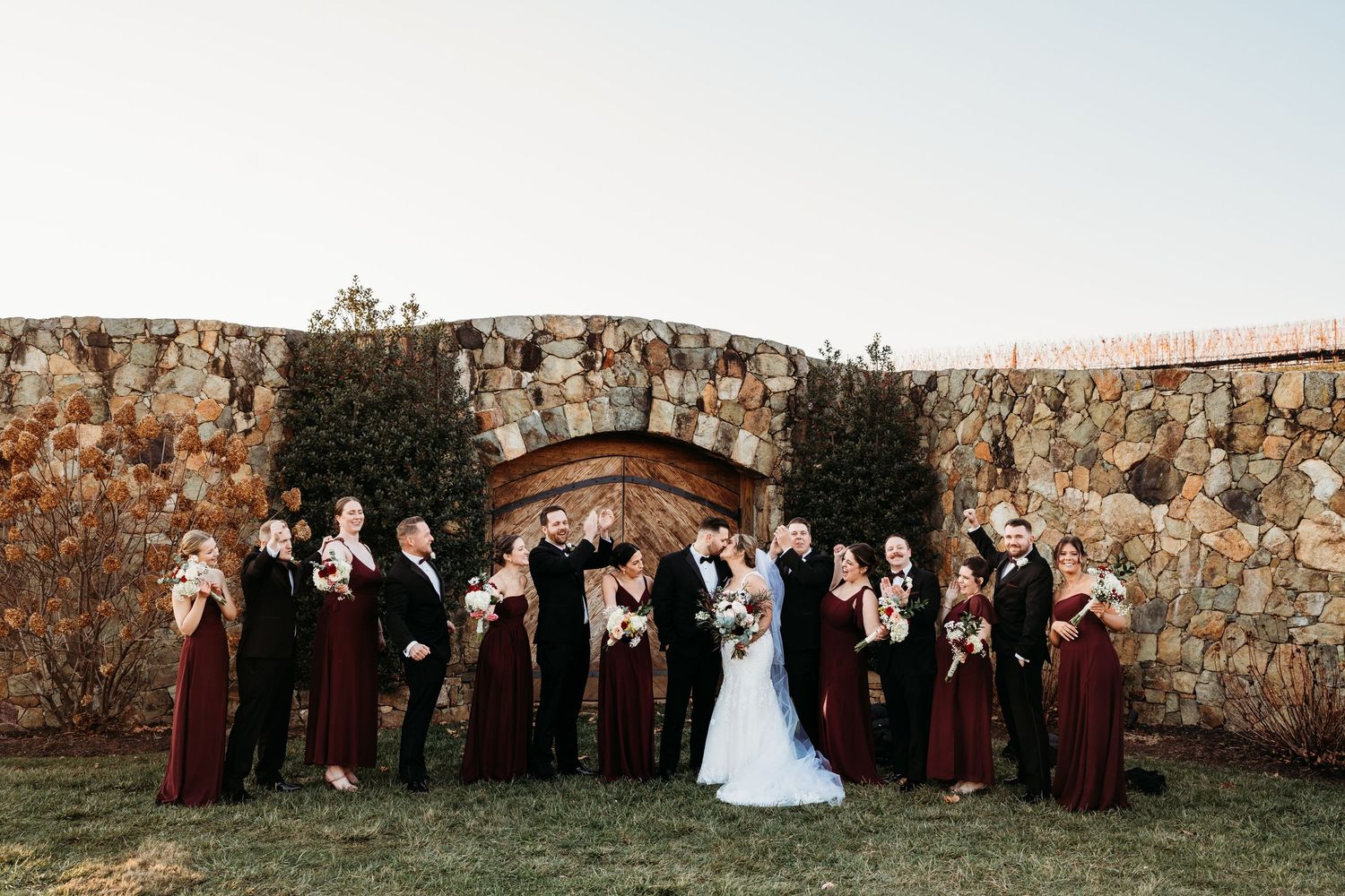 Wedding party lines up against stone wall for formal group photo at sunset.