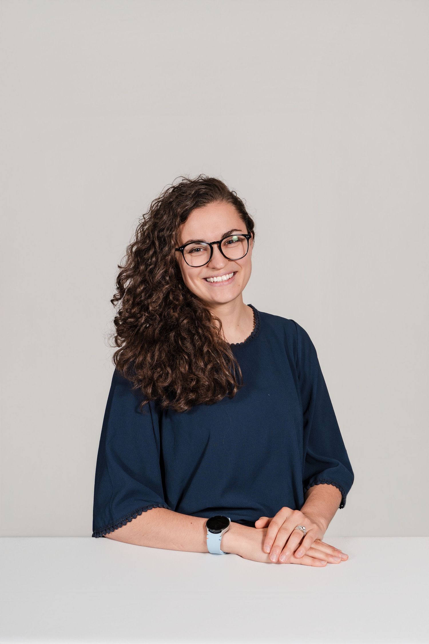 Person with curly dark hair and glasses wearing a navy blue shirt poses at a white desk in a photo series.