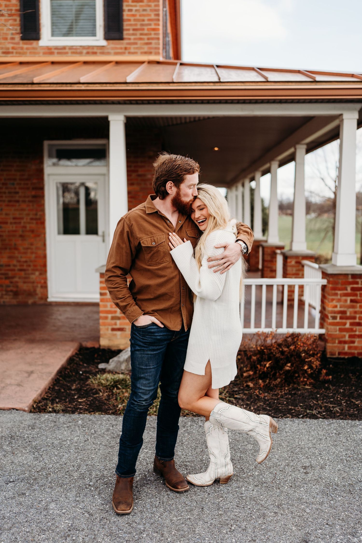 A couple embracing on a driveway in front of a red brick house with white trim.