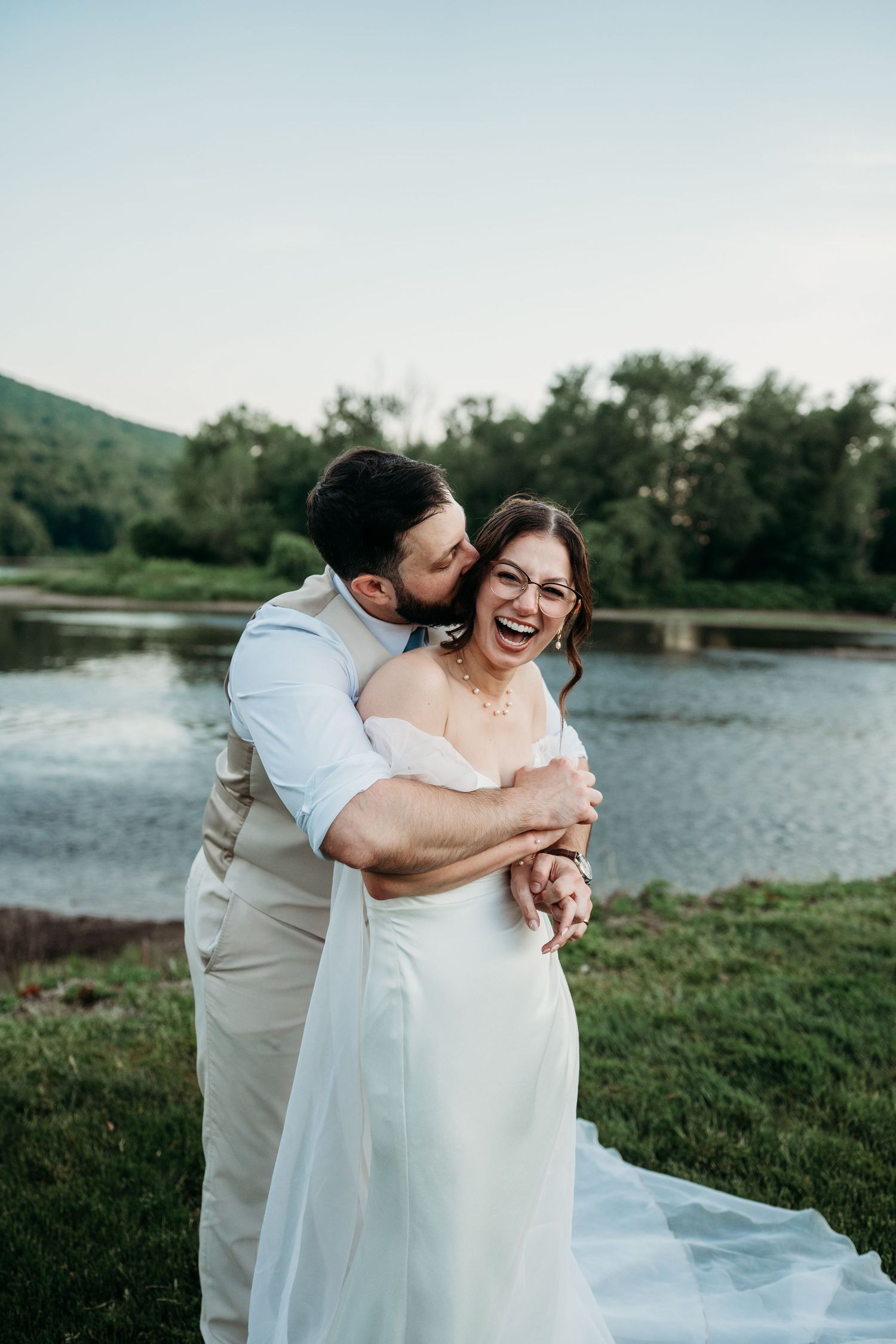 Joyful bride in white off-shoulder gown laughs as groom embraces her from behind near a serene lake.