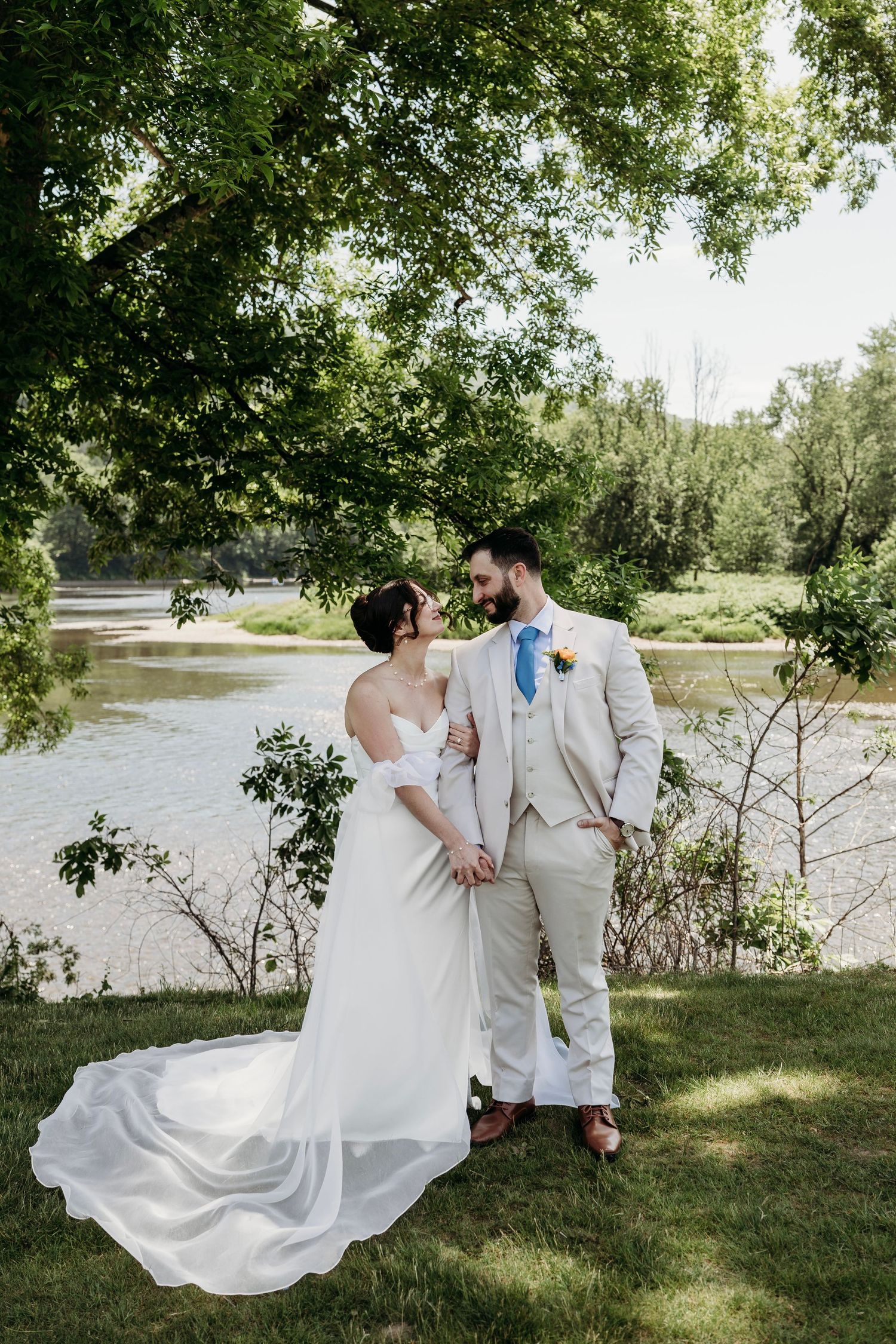 Bride and groom hold hands by a sunlit river, surrounded by lush green trees, as she wears a flowing white gown.