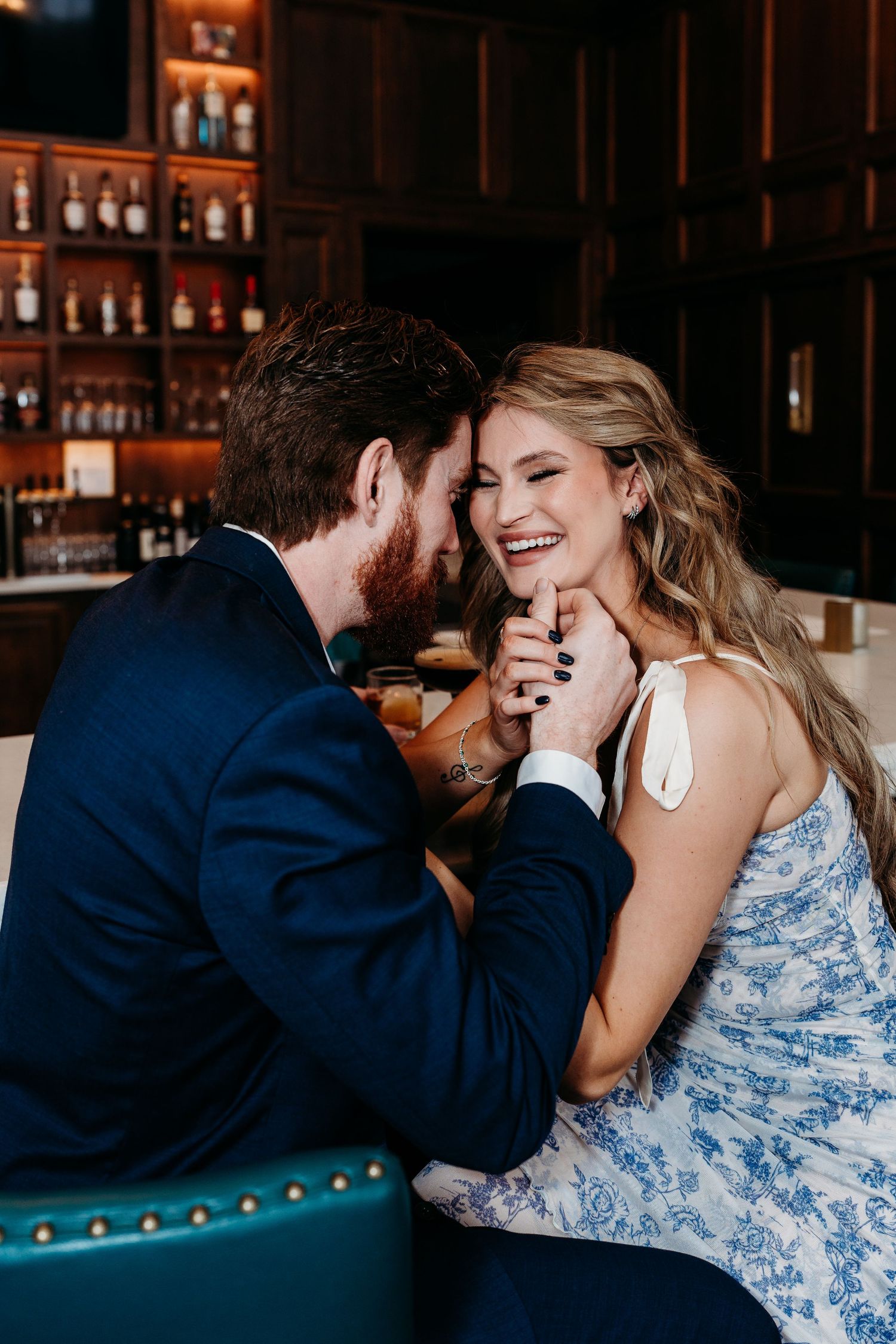 A couple shares an intimate moment at a bar with blue seats and bottles displayed in the background.